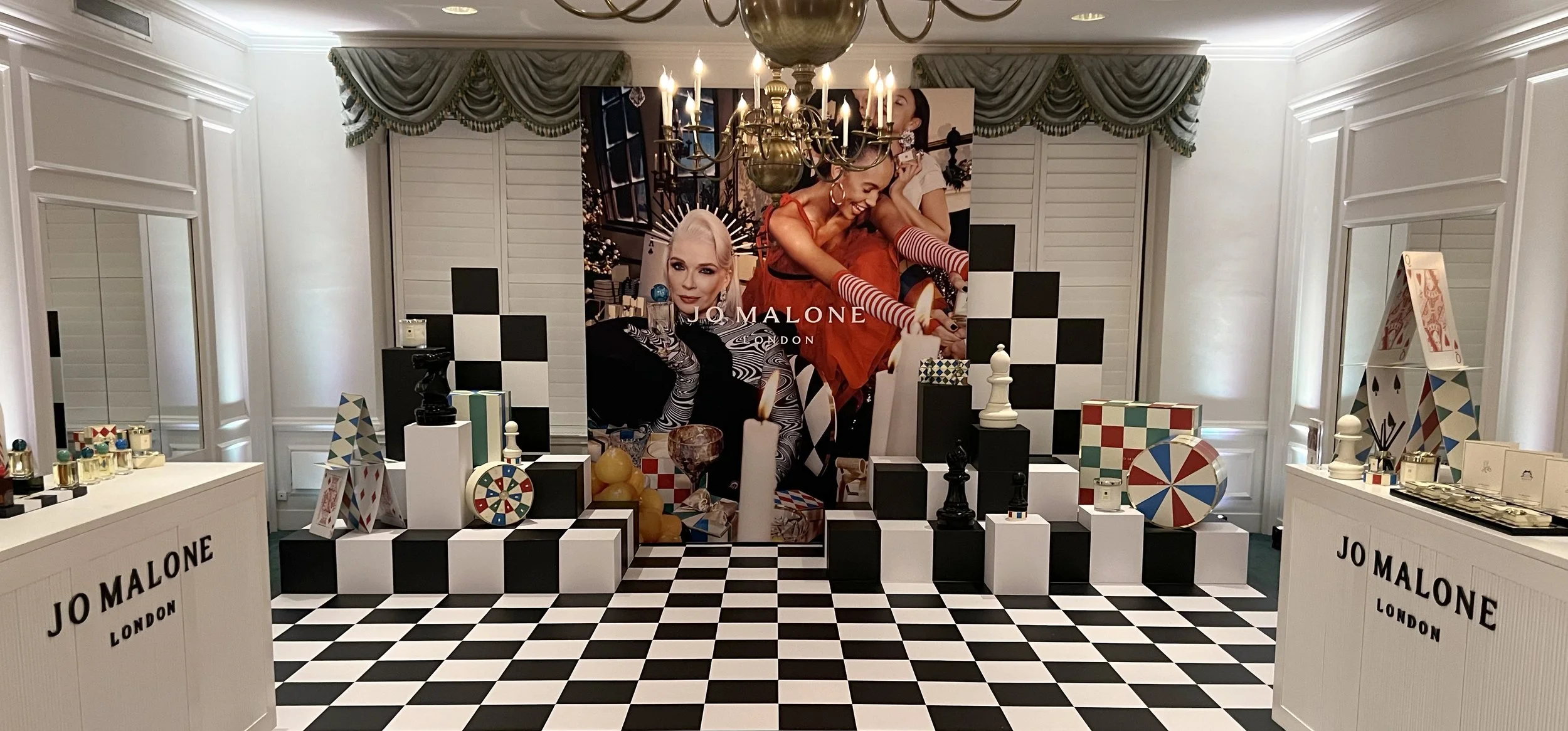 Jo Malone London display with black and white checkered flooring, black and white squares on the back wall, and a large advertisement featuring a smiling woman and a girl. The display includes colorful decorative objects, candles, and a chess piece, 