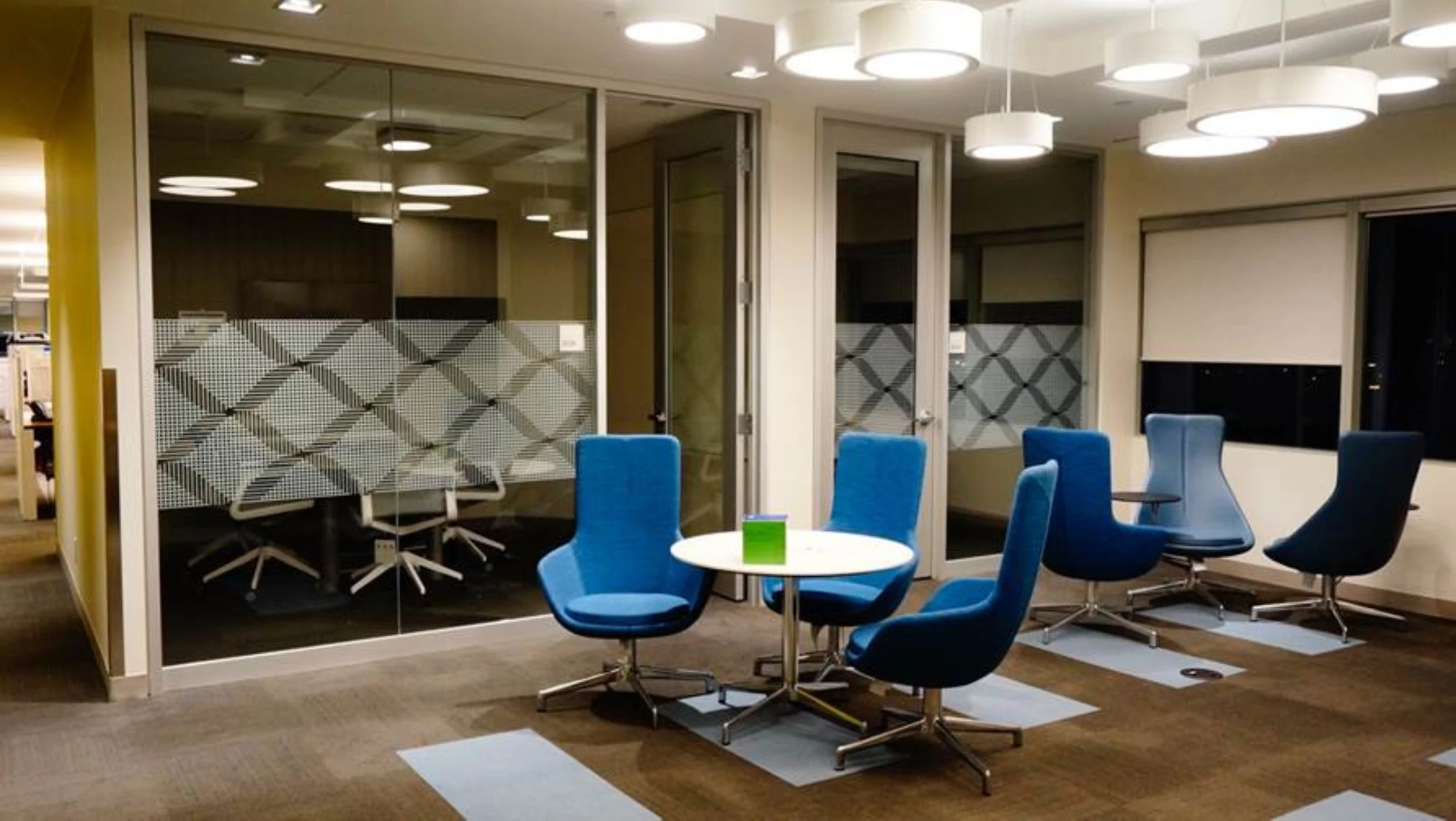 Modern office meeting room with blue chairs, a white round table, and a glass-walled conference room in the background.