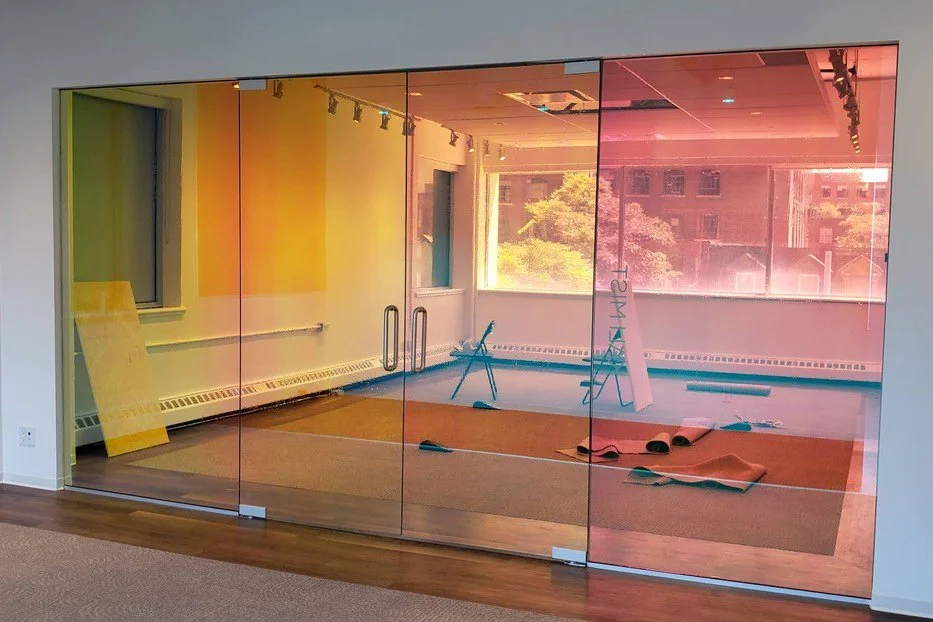 ~A glass wall with rainbow-like tinted glass separating an office space from a room with large windows, mats, a ladder, and equipment inside.