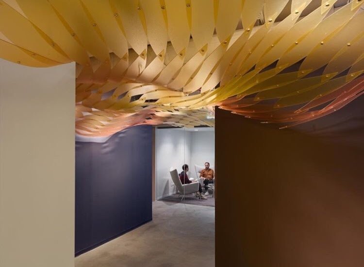 ~Interior view of a modern office or lounge with a decorative yellow and orange ceiling installation, dark and light walls, and two people seated in conversation in a seating area.