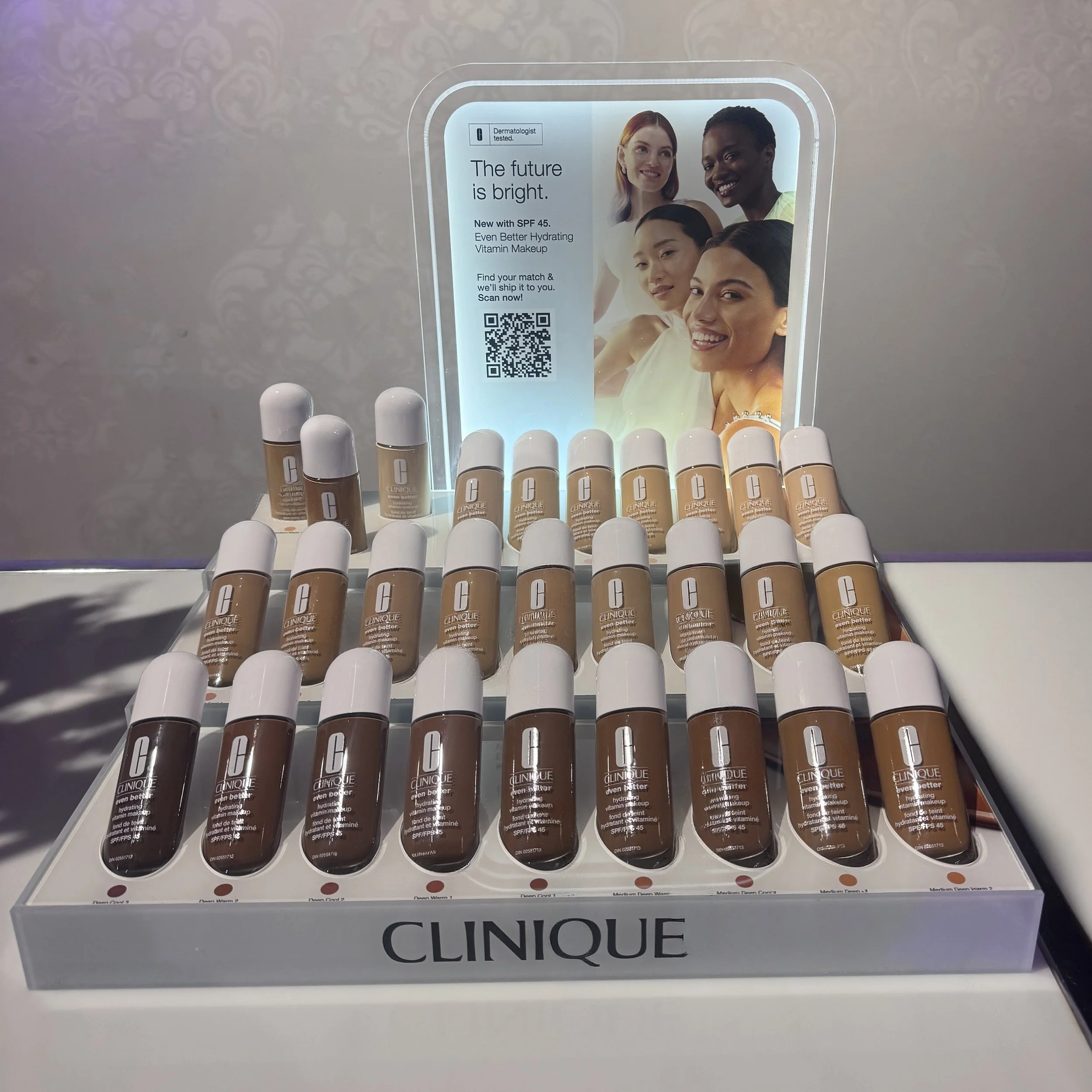 Display of Clinique foundations in varying shades with a promotional signage in the background featuring diverse women smiling.