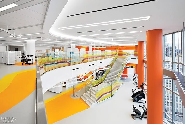 Modern interior of a multi-level commercial or office space with orange accents, glass railing, stairs, exercise equipment, and large windows showing city buildings.