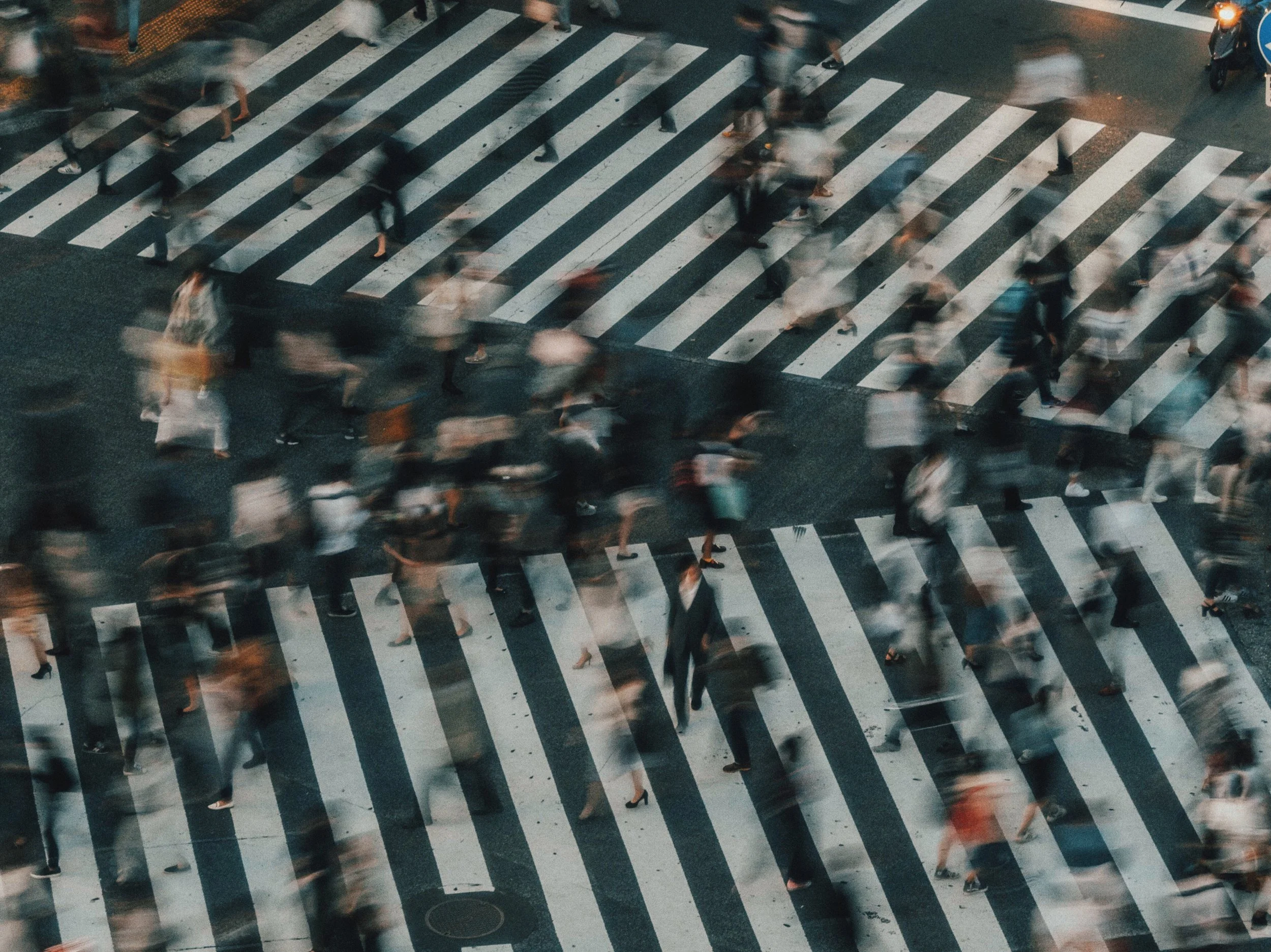 Aerial view of a busy crosswalk with many blurred pedestrians walking in different directions, captured during evening or nighttime.