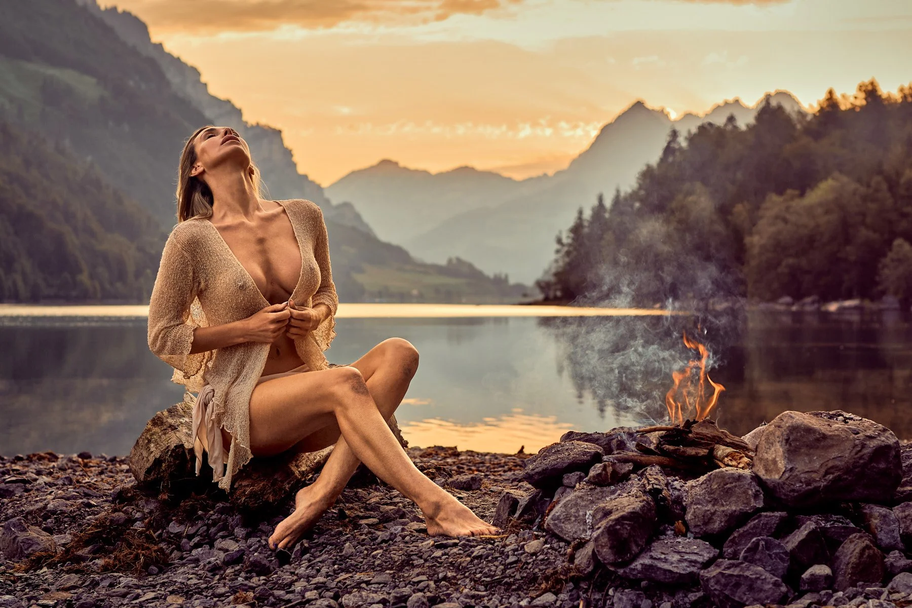 Eine Frau sitzt am Ufer eines Flusses bei Sonnenuntergang, mit einem kleinen Feuer vor ihr, umgeben von Bergen und Bäumen.