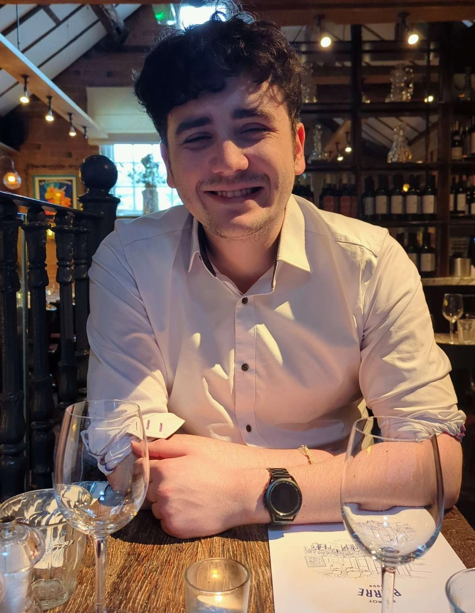 A smiling man with dark, curly hair wearing a white shirt sitting at a restaurant table with glassware and a menu, inside a cozy restaurant with wooden walls, shelves with wine bottles, and warm lighting.