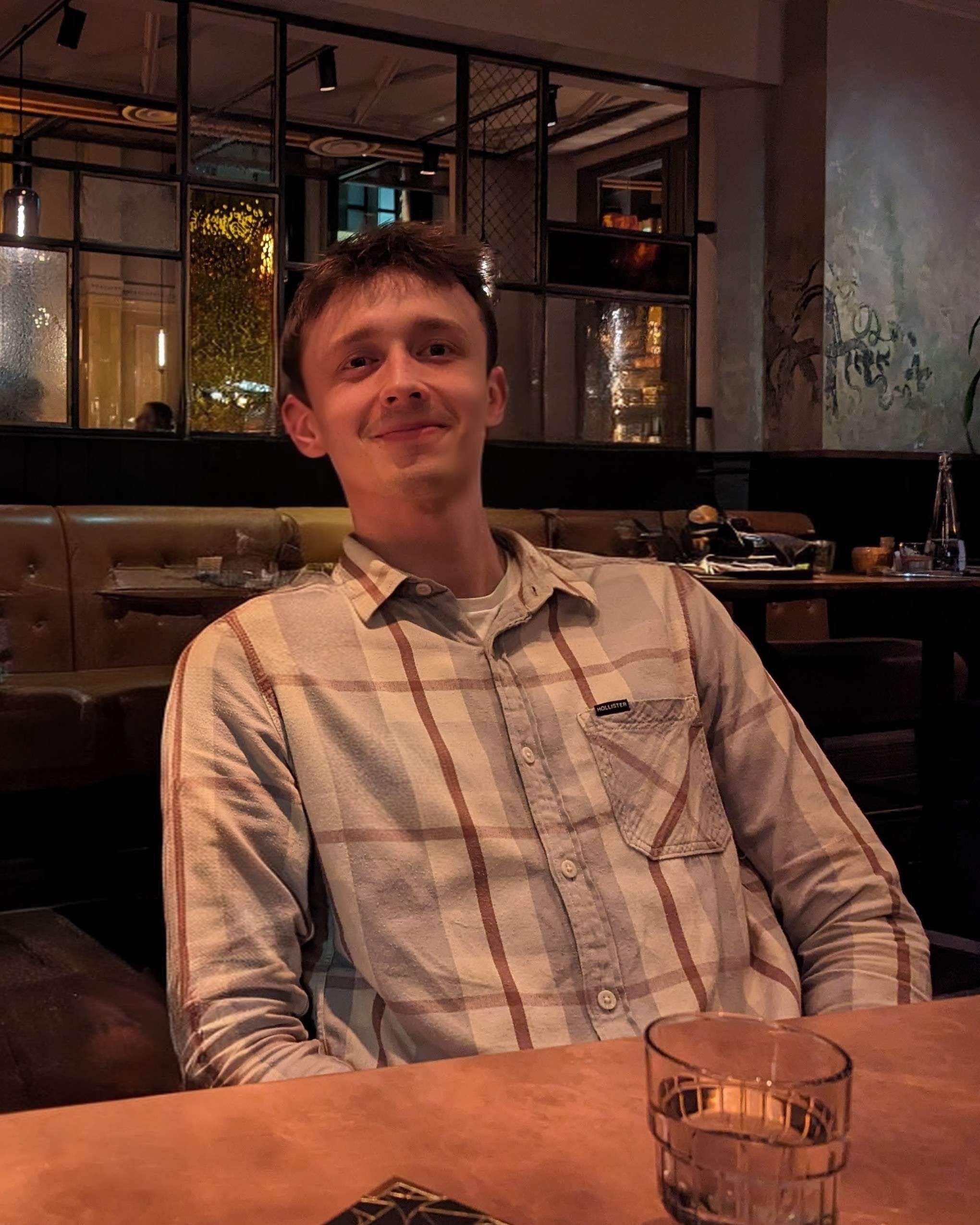 A young man sitting at a restaurant table smiling at the camera, wearing a plaid shirt. The restaurant has dark wood and leather decor with warm lighting.