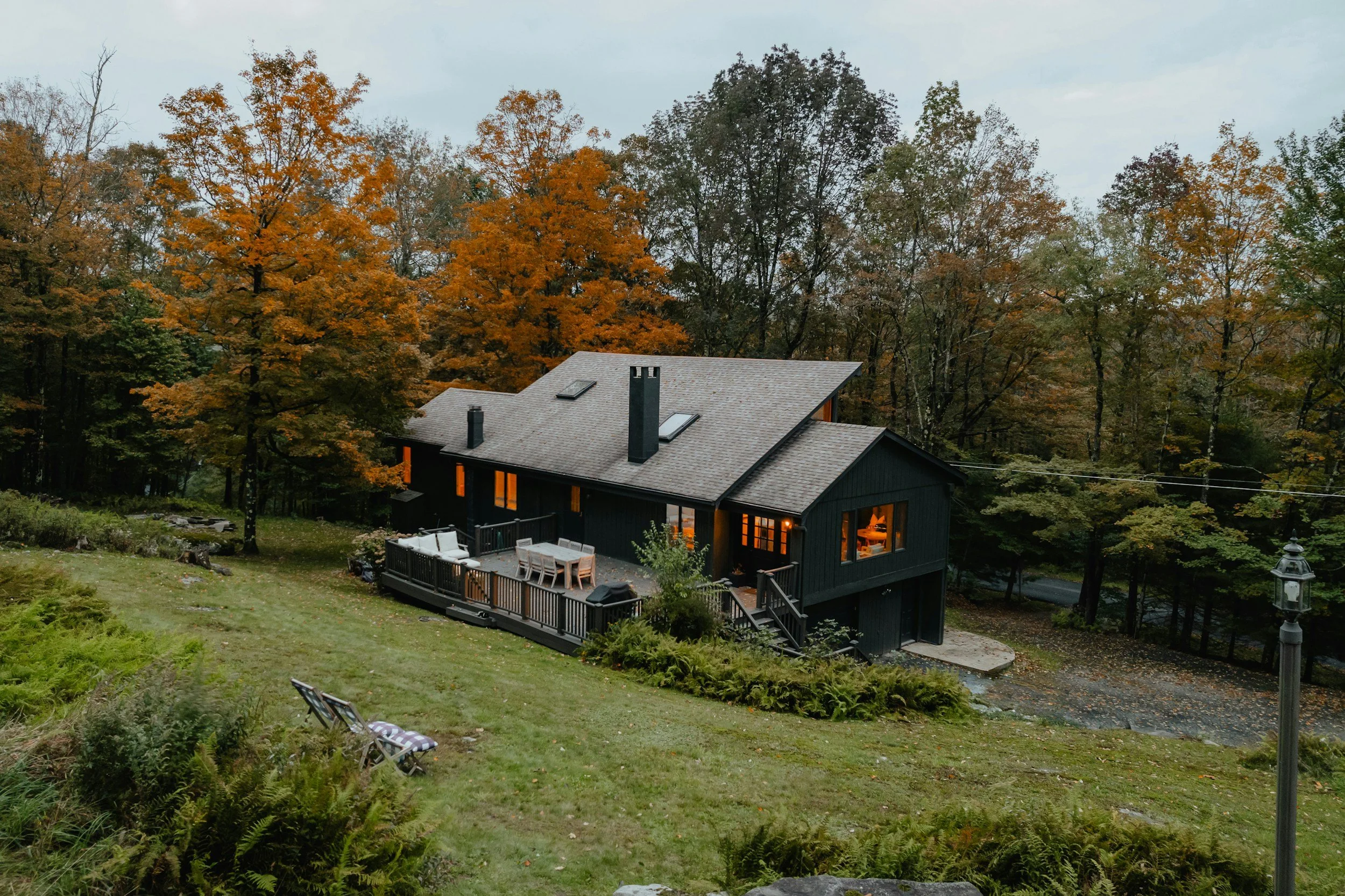 Black house with illuminated windows, wooden deck, and outdoor furniture, surrounded by autumn trees with orange and green leaves, on a sloped grassy yard.