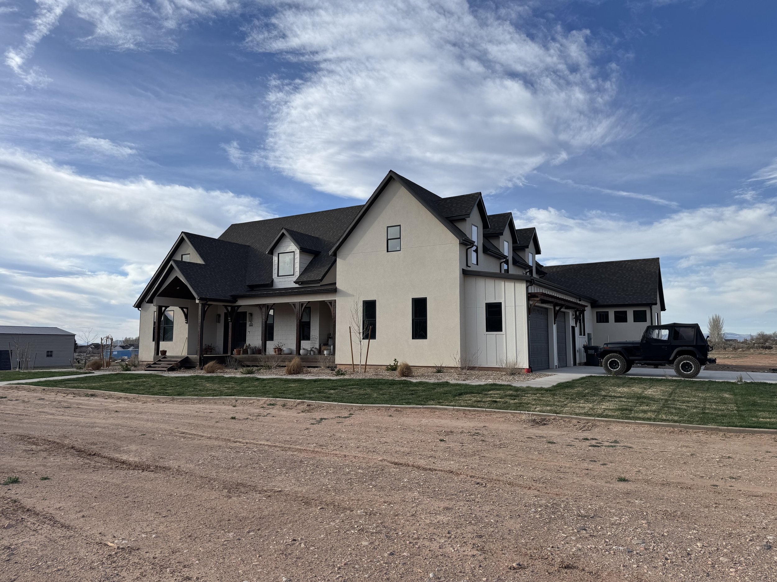 Modern house with gray siding and black roof, front porch, yard with grass and mulch, driveway with black Jeep, and cloudy sky