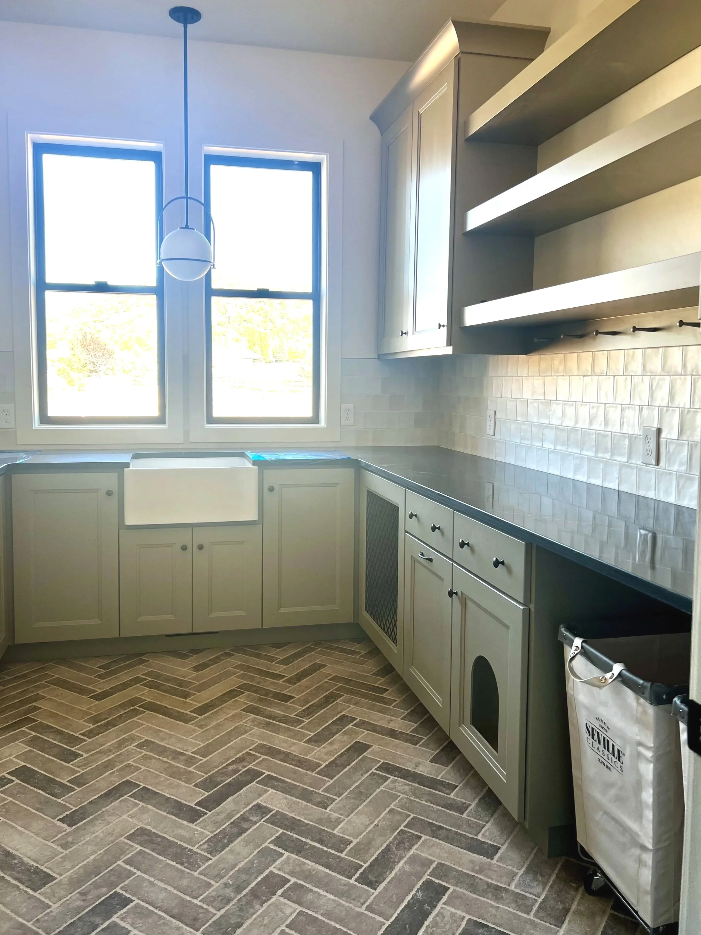 Laundry room with sage cabinets, a white farmhouse sink, a dark soapstone countertop, open shelves, and two large windows to let in the sunlight. Notice pet-focused specialty lower cabinets.