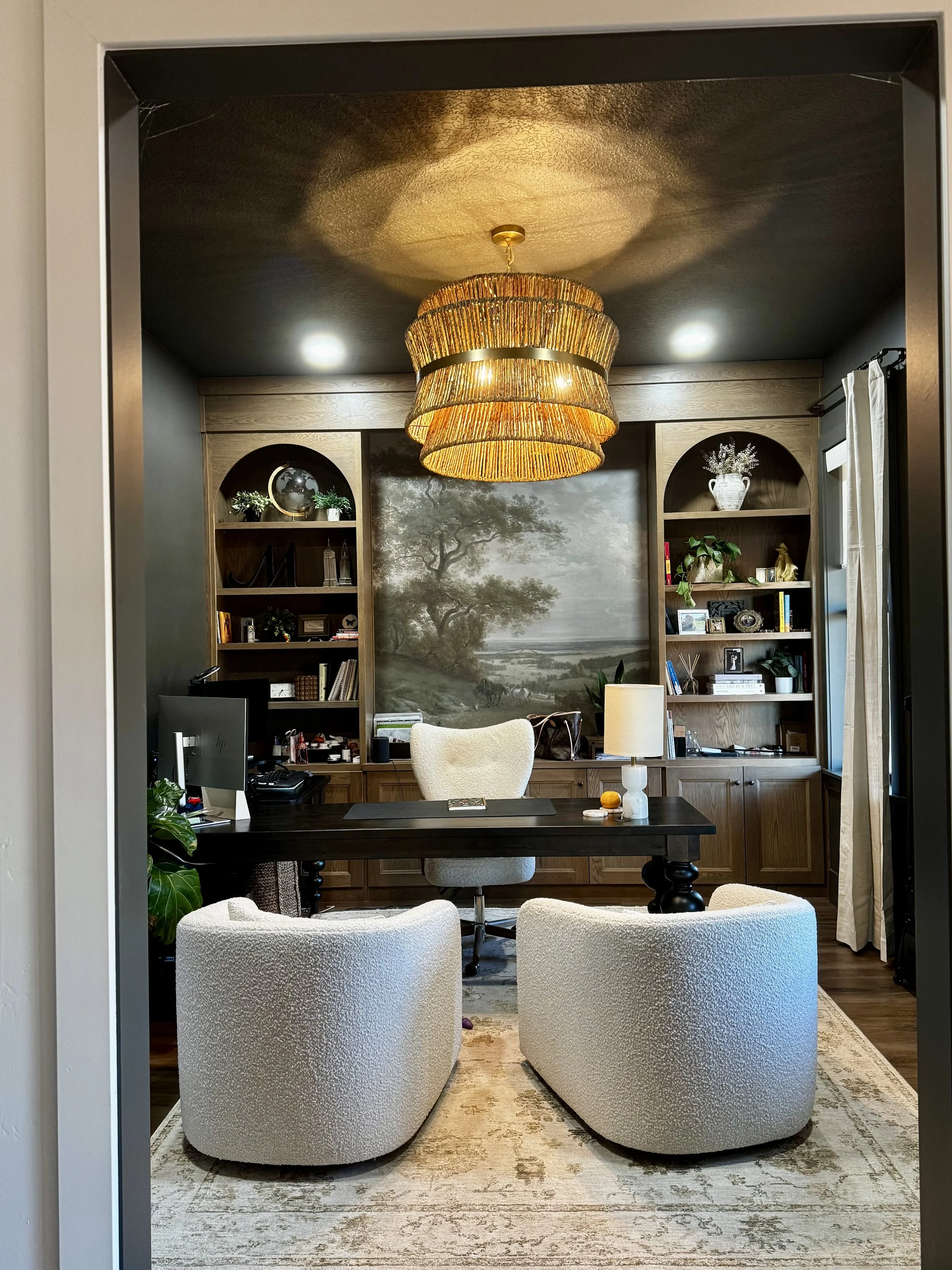A home office with a dark wood desk, cream armchairs, and built-in wooden bookshelves behind. A large, woven chandelier hangs from the ceiling, illuminating the room. The back wall features a mural of a serene landscape with a tree and river.