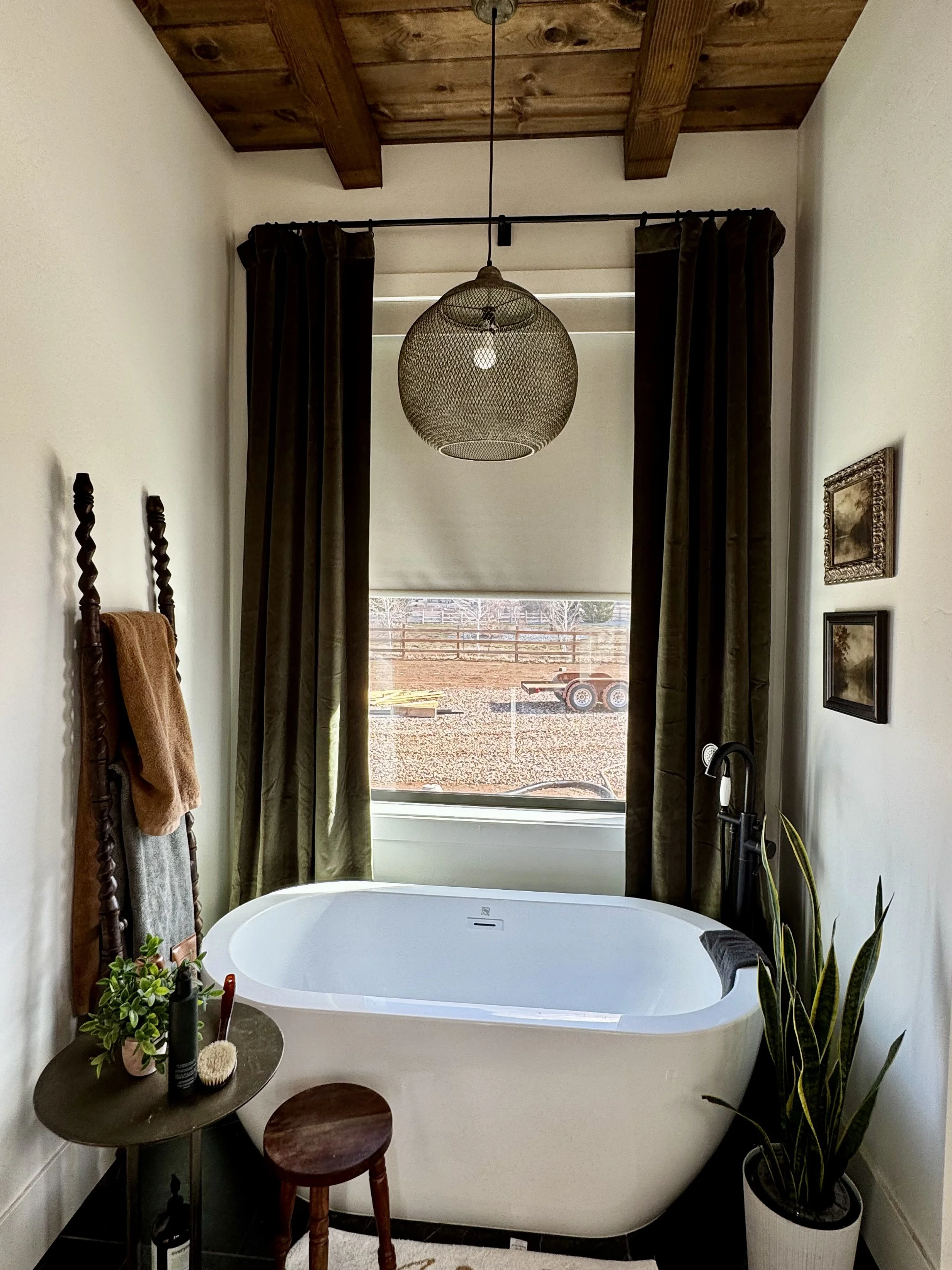 A cozy bathroom featuring a white bathtub beneath a window with dark green curtains. There is a wooden ladder-style towel rack with towels, a small round side table with plants and toiletries, a tall plant in a white pot, and decorative framed pictures on the wall. A woven pendant light hangs from a wooden beam ceiling.