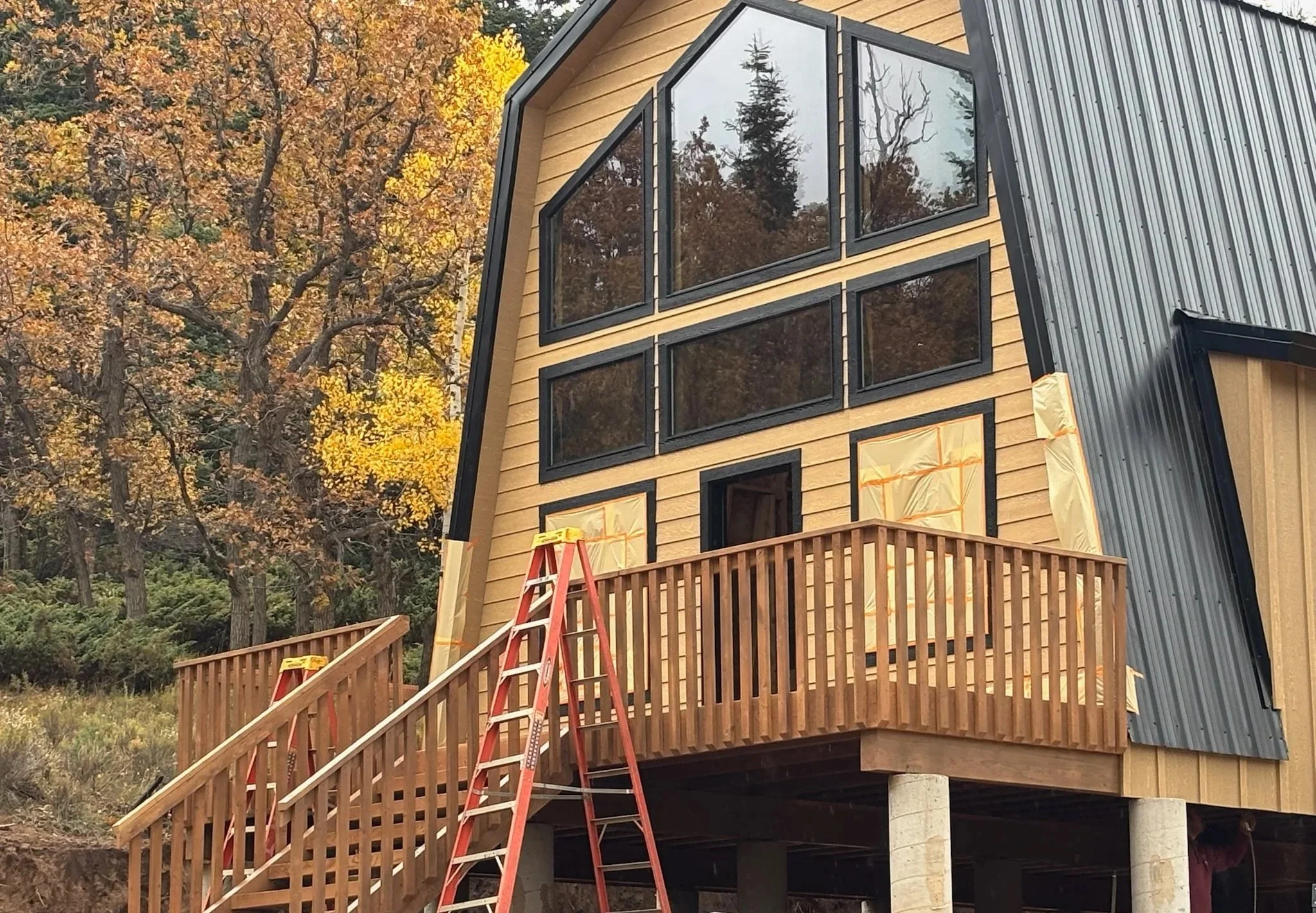 A modern house under construction with a wooden and metal exterior, large windows, a wooden balcony, and a red ladder, surrounded by trees with fall foliage.