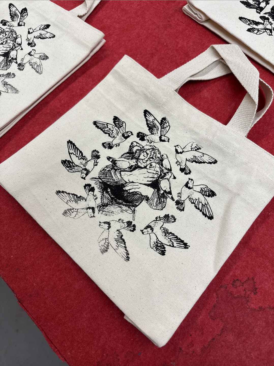 A white tote bag with a black illustration of a person hugging a pigeon surrounded by flying pigeons, on a red surface.
