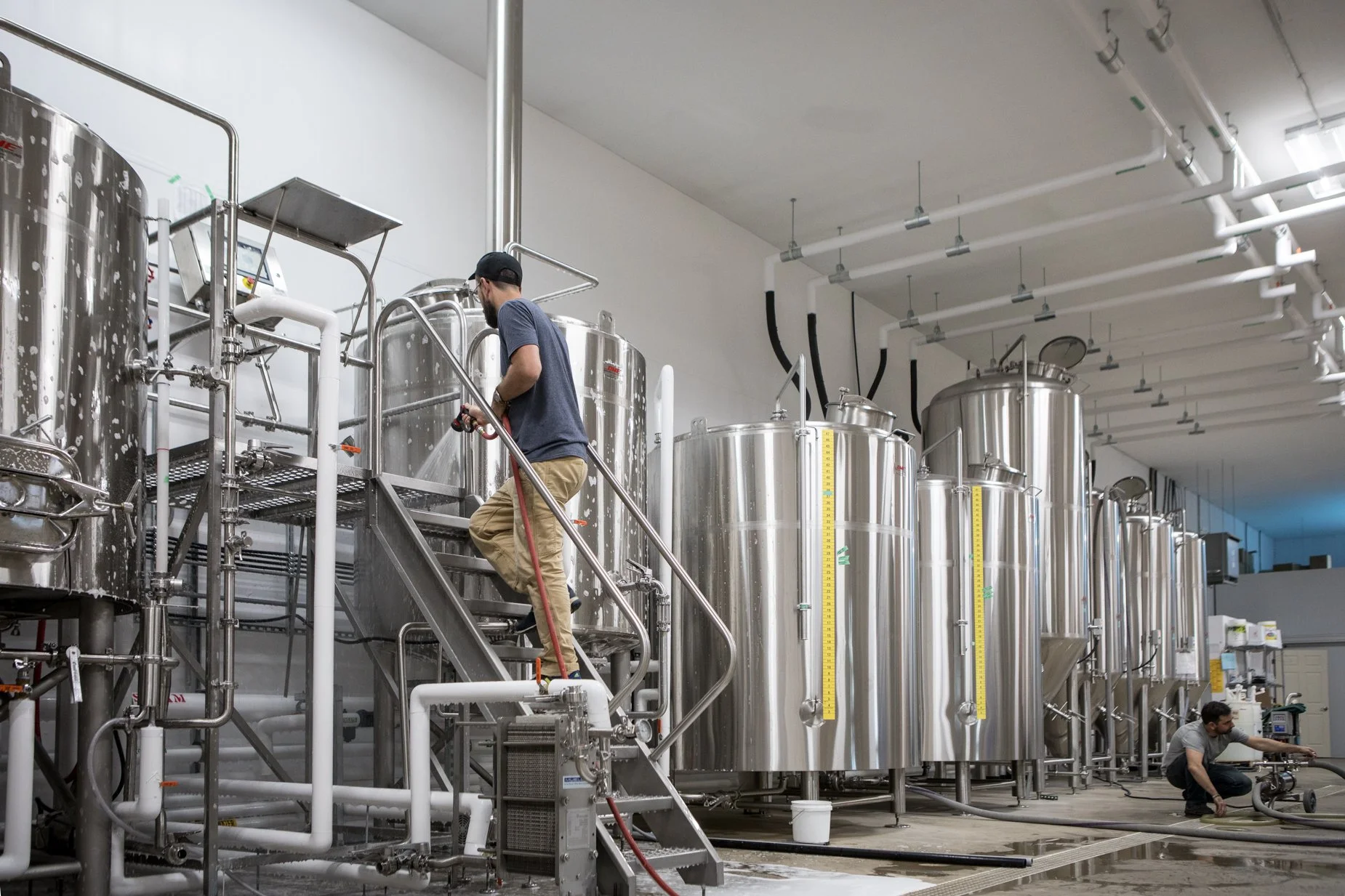 Two workers in a brewery with stainless steel fermentation tanks, one washing a tank and the other operating equipment, inside a modern brewery factory.