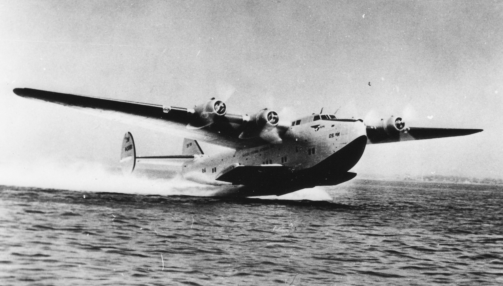 Black and white photo of a large seaplane with four engines taking off from water, creating a wake behind it. The sea is visible beneath the plane, and the sky appears overcast.