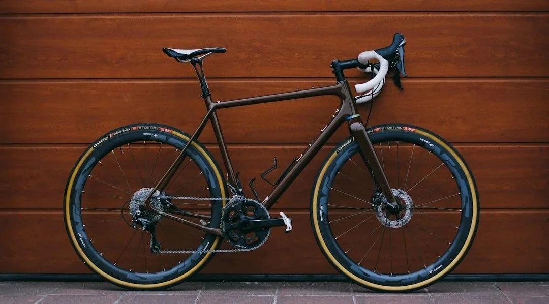 A modern black and brown gravel bike with white handlebars and yellow accents on the tires, mounted against a wooden wall.
