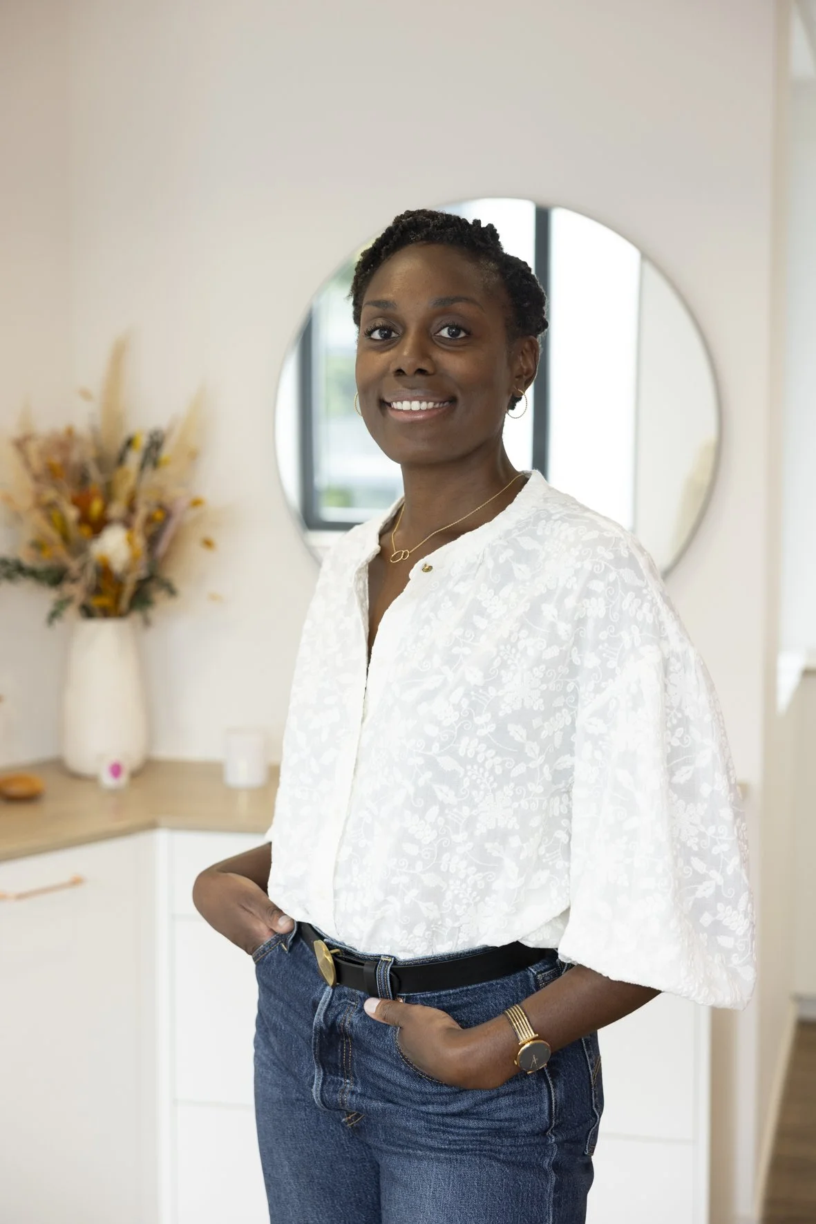 A woman with short curly hair wearing a white blouse, blue jeans, and accessories, standing indoors with a mirror and a floral arrangement in the background.