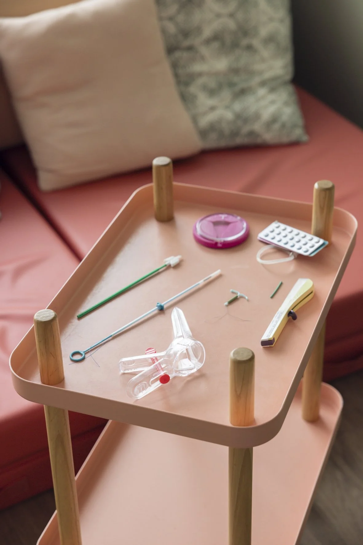 Une table rose claire avec des instruments médicaux pour l'autoscopie et des tubes à essai, placée devant un canapé avec deux coussins, un beige et un gris. Suivi gynécologique de prévention ,contraception, frottis à Rennes Cesson-Sévigné. Chalotais