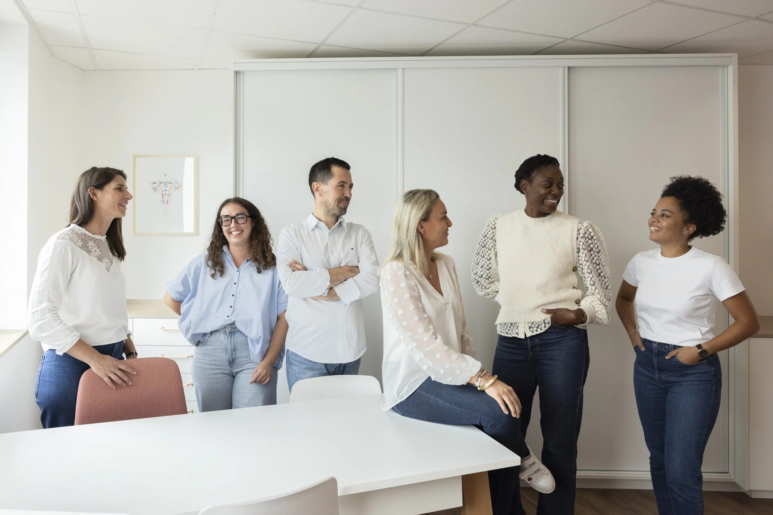 Six women and one man standing and sitting in a bright office, smiling and engaging in conversation.