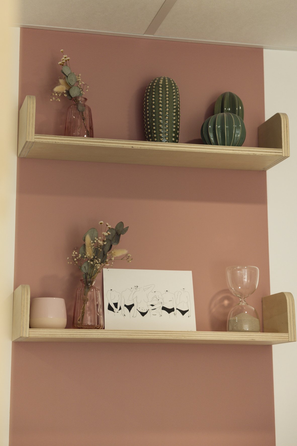 Decorative wooden shelves on a pink wall with vases, cactus-shaped objects, a drawing of nude figures, a pink vase with flowers, a pink candle holder, and an hourglass.