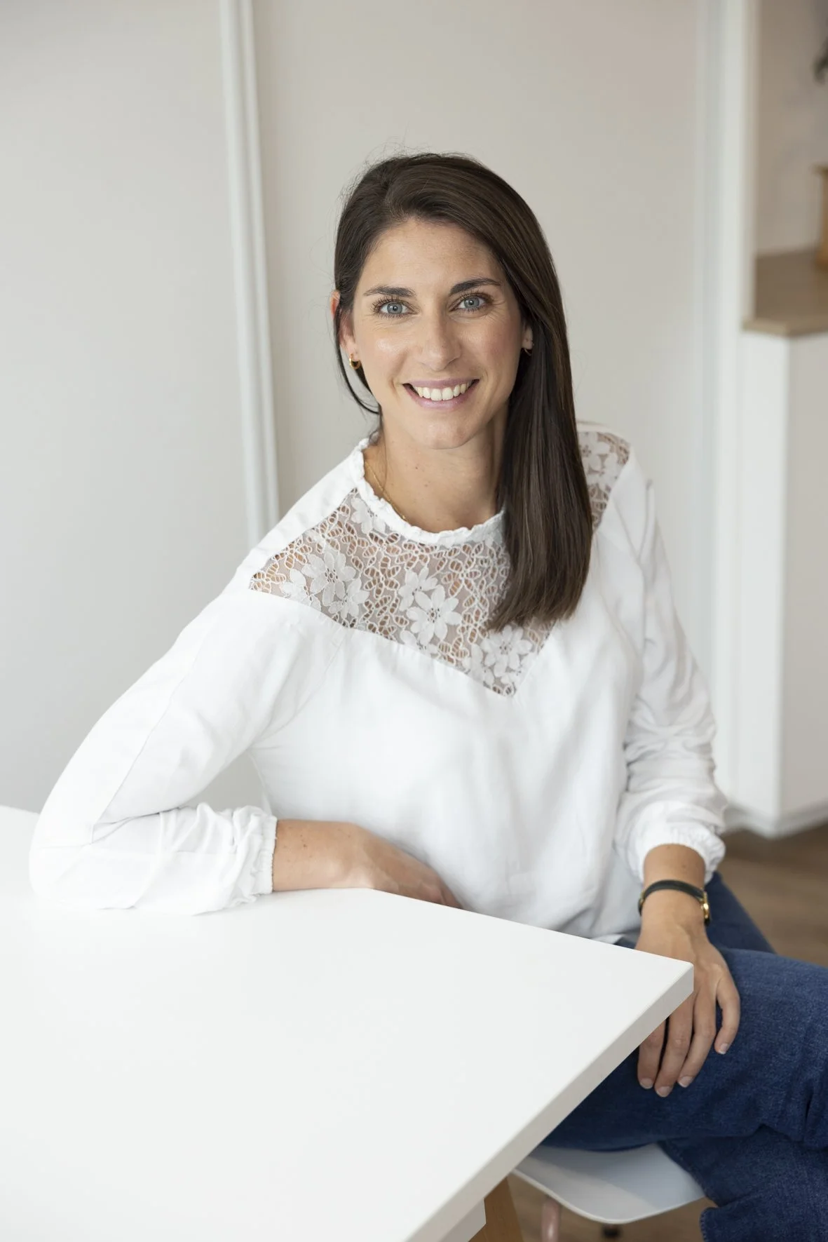 Femme souriante assise, portant une chemise blanche avec détails en dentelle sur le haut, dans une pièce au décor minimaliste. Eve GALLOUX Sage-Femme au Cabinet Chalotais à Cesson-Sévigné