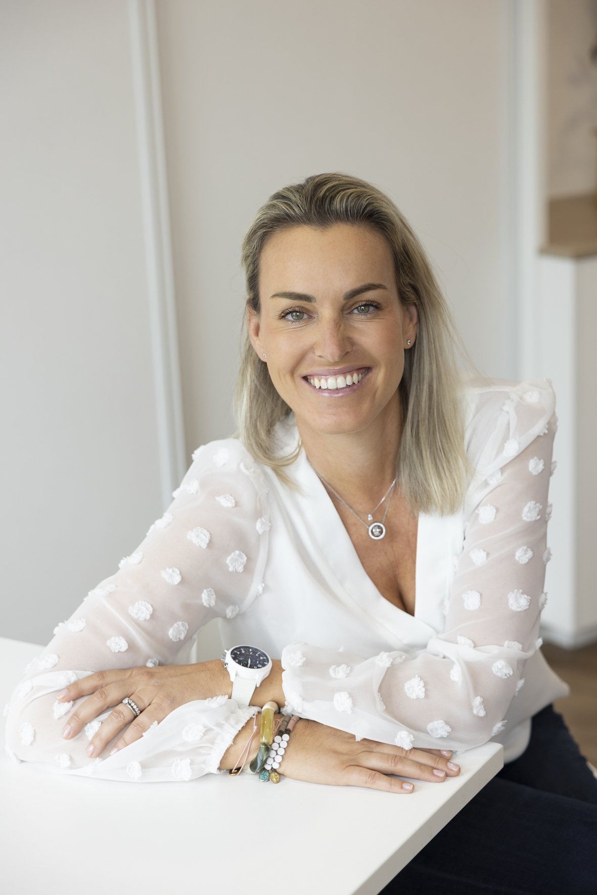 Une femme souriante avec des cheveux blonds, portant une chemise blanche à pois en relief, assise à une table blanche dans un intérieur lumineux, portant une montre blanche et des bracelets. Svenja DELACOUR ISLER Sage-Femme au Cabinet Chalotais