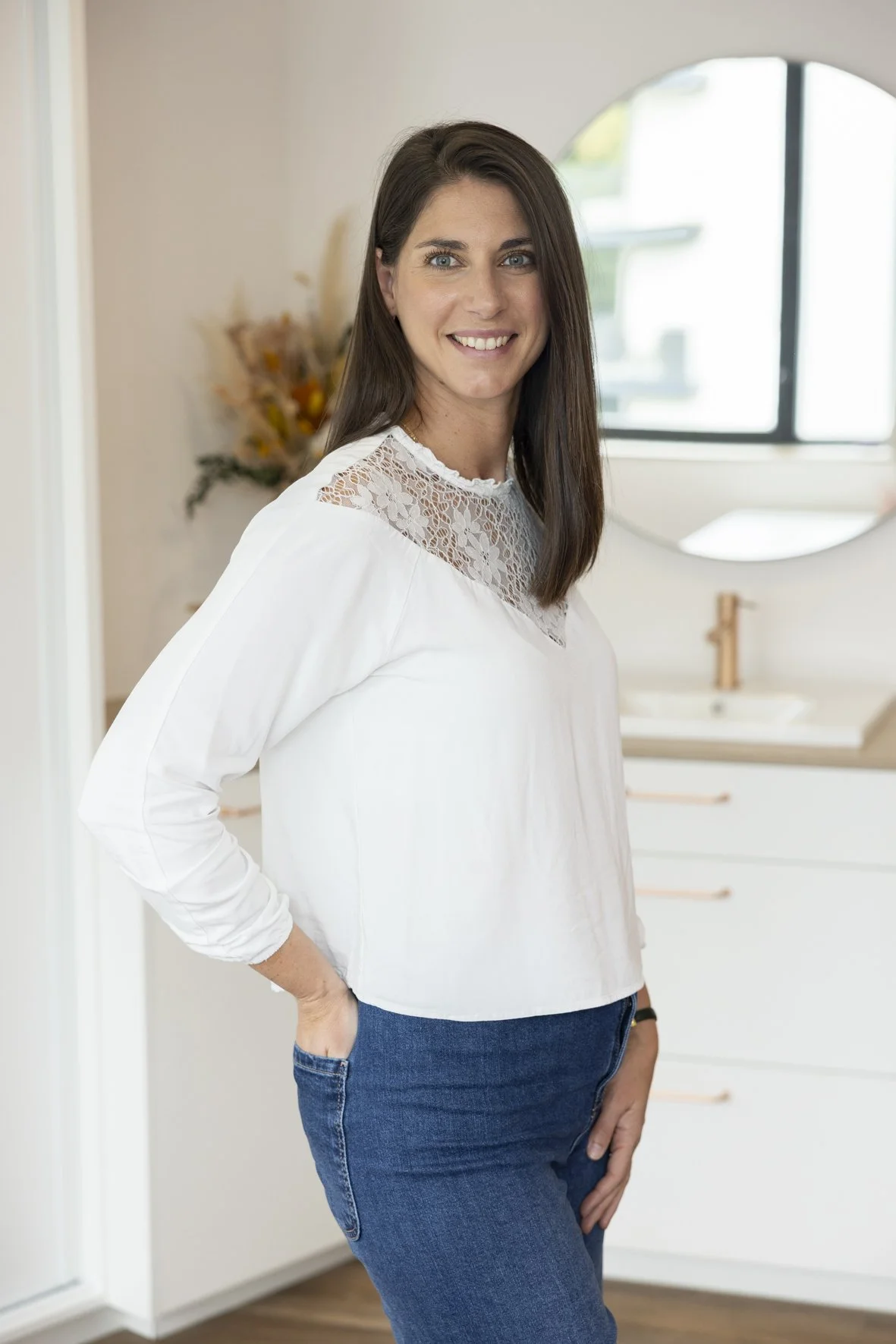 Smiling woman with long brown hair, wearing a white long-sleeve shirt with lace details, standing in a bright room with a bathroom mirror and sink in the background.