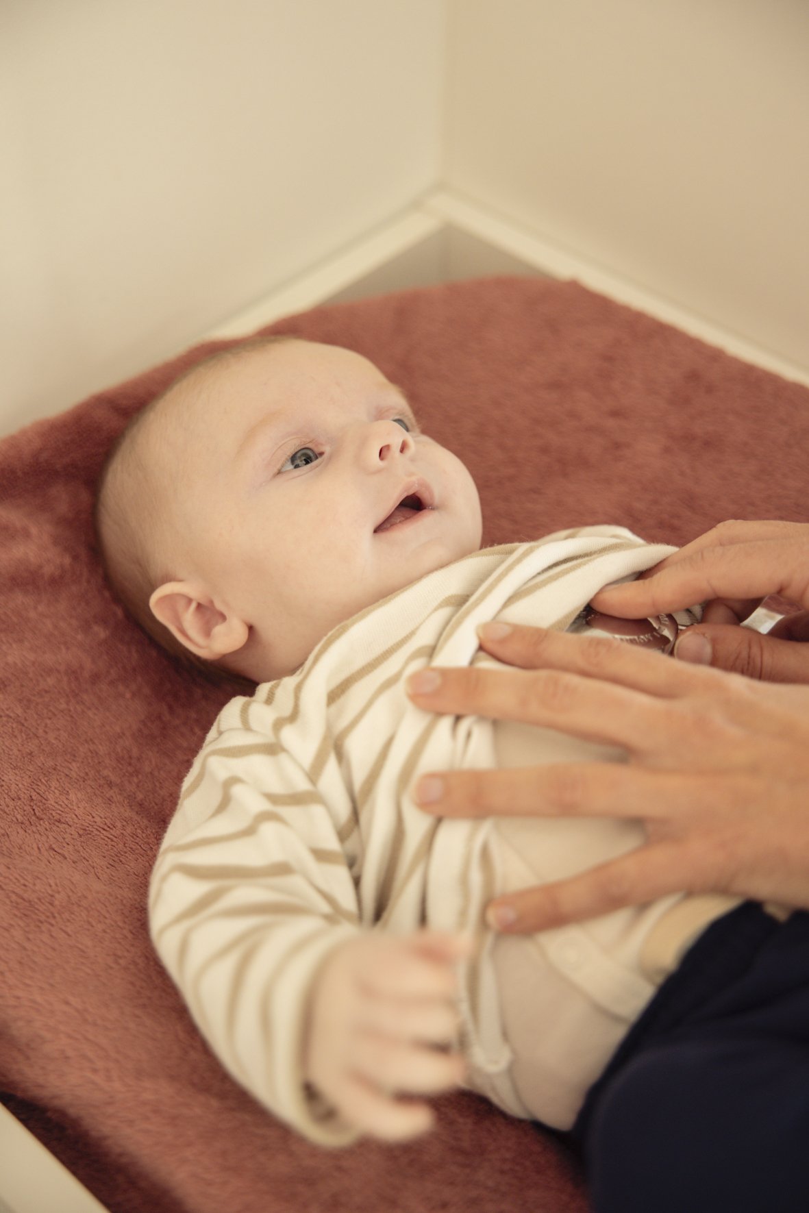 A baby lying on a pink and brown surface, looking up with an open mouth, while an adult's hands gently hold the baby's chest.