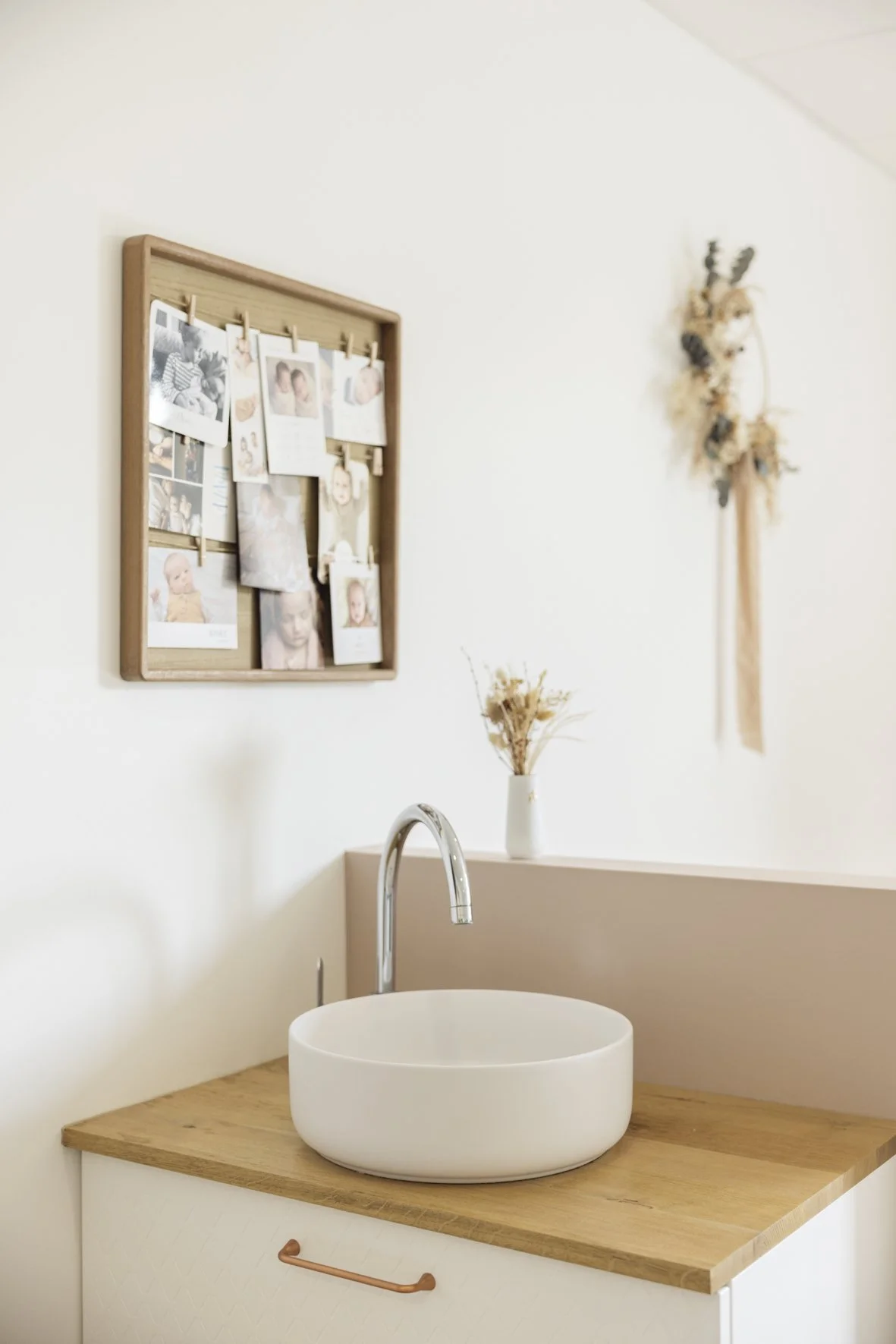 Bathroom with a round white vessel sink, wooden countertop, brass drawer handle, small vase with dried flowers, photo collage on the wall, and dried floral wall hanging.