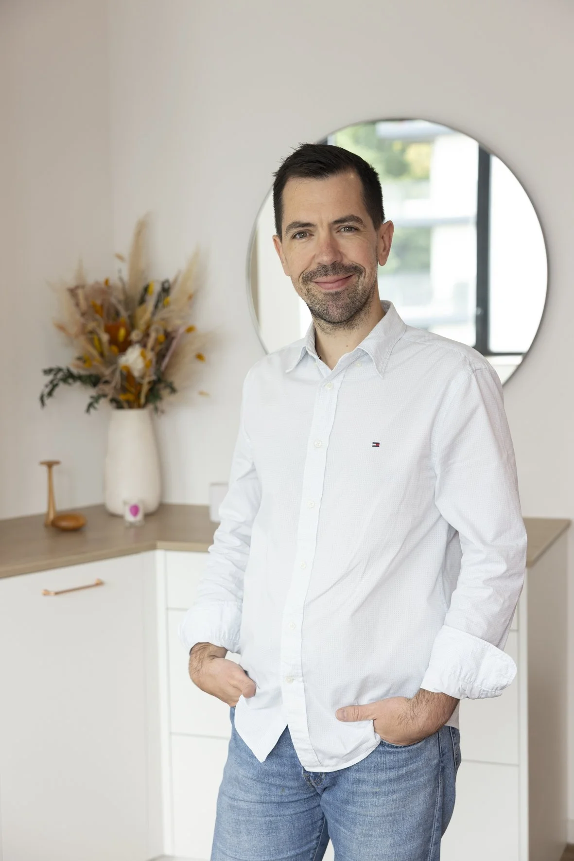 A man with dark hair and a beard wearing a white button-up shirt and jeans, standing indoors with hands in pockets, in front of a wall with a circular mirror and decorative vase with flowers.