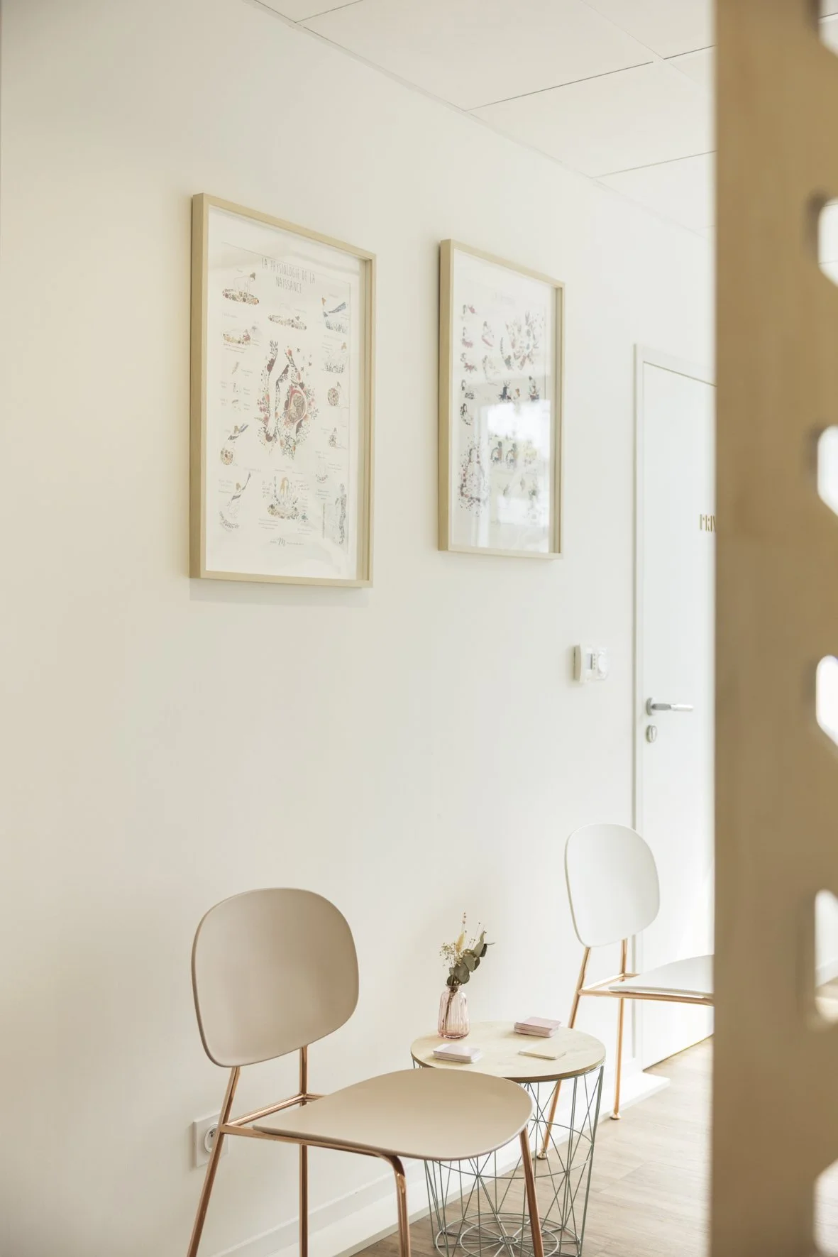 Une salle d'attente minimaliste avec deux chaises blanches en métal, une petite table ronde avec un vase contenant des fleurs, et deux cadres suspendus contenant des illustrations sur le mur blanc. Suivi post partum Rennes. Cabinet Chalotais Cesson