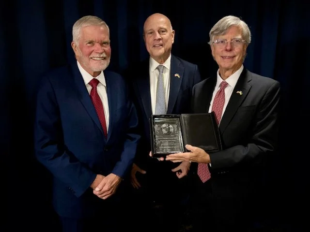 Three men in suits standing in front of a dark blue curtain, with the man on the right holding a clear glass award or plaque in a case.