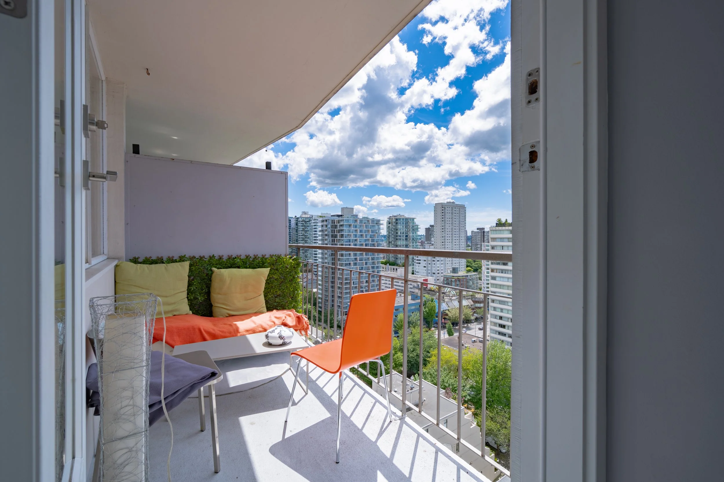 Balcony with seating area, including pillows and chairs, overlooking cityscape with tall buildings under a partly cloudy sky. Vancouver BC, 1903 Comox St.