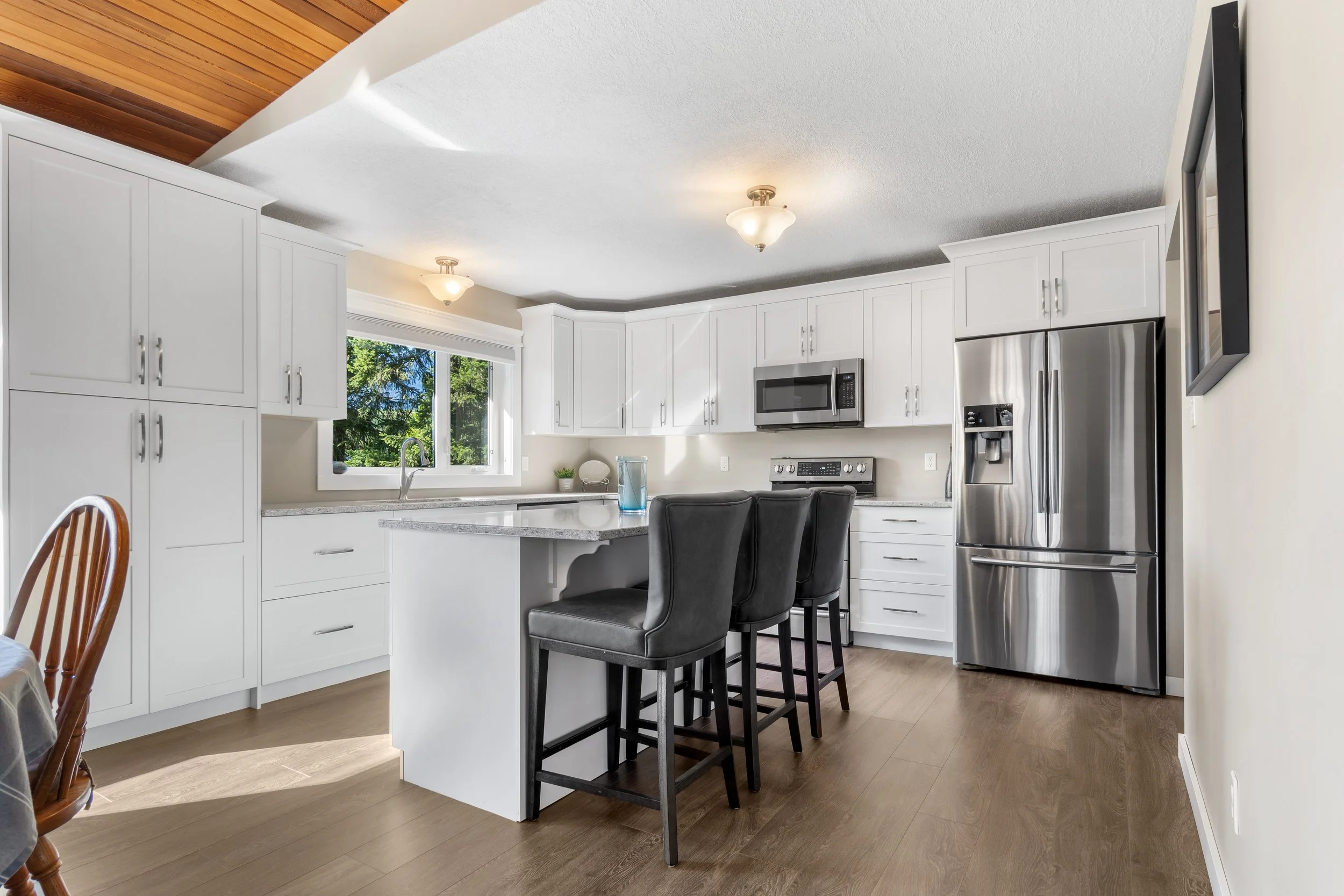 Modern white kitchen with wood flooring, stainless steel appliances, black barstools at kitchen island, large window showing green trees, with a wooden ceiling detail and bright lighting.