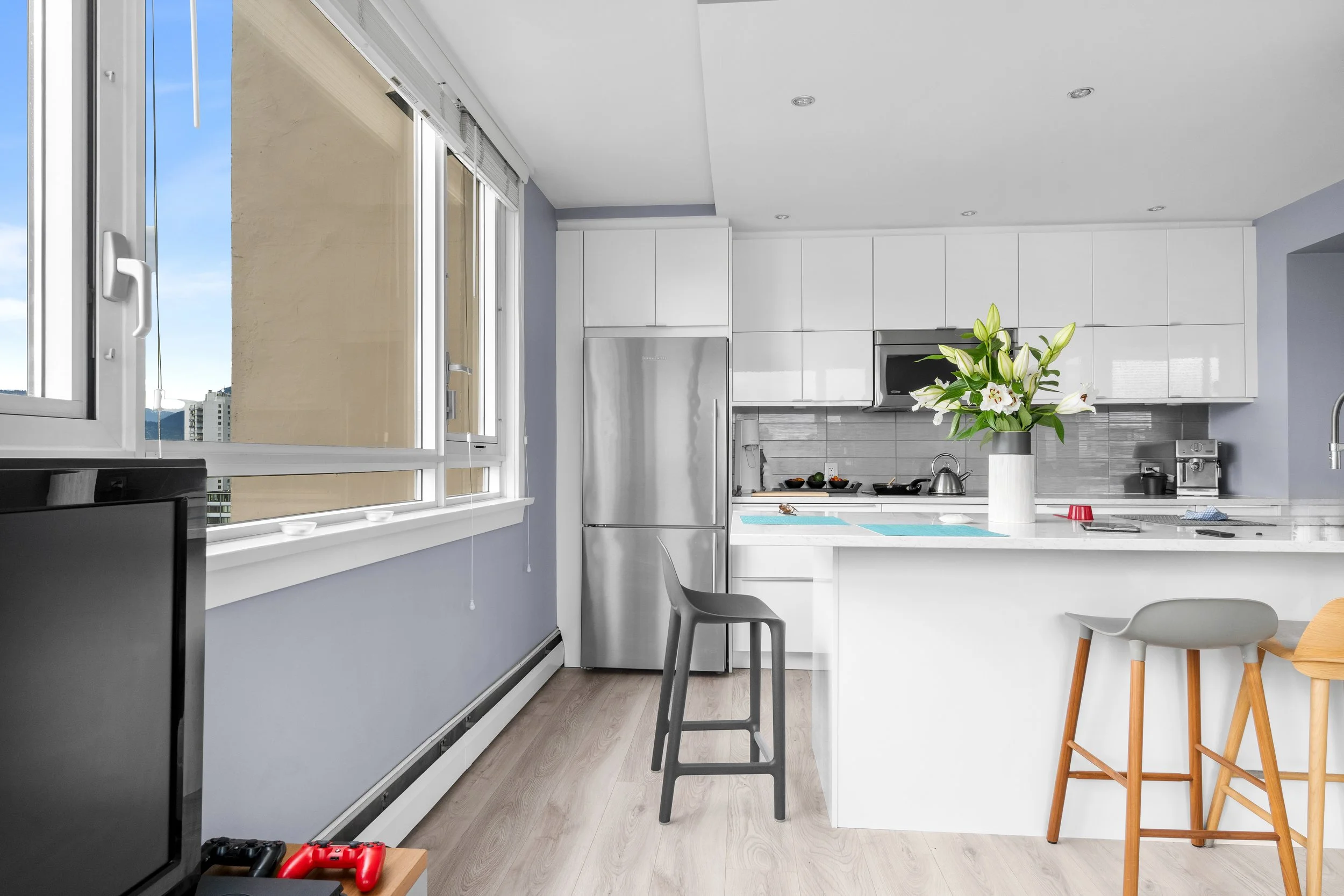 Modern kitchen with light gray walls, white cabinets, stainless steel appliances, and a large window with a city view. There is a vase of white lilies on the kitchen island, and seating includes a gray chair and wooden high stools.