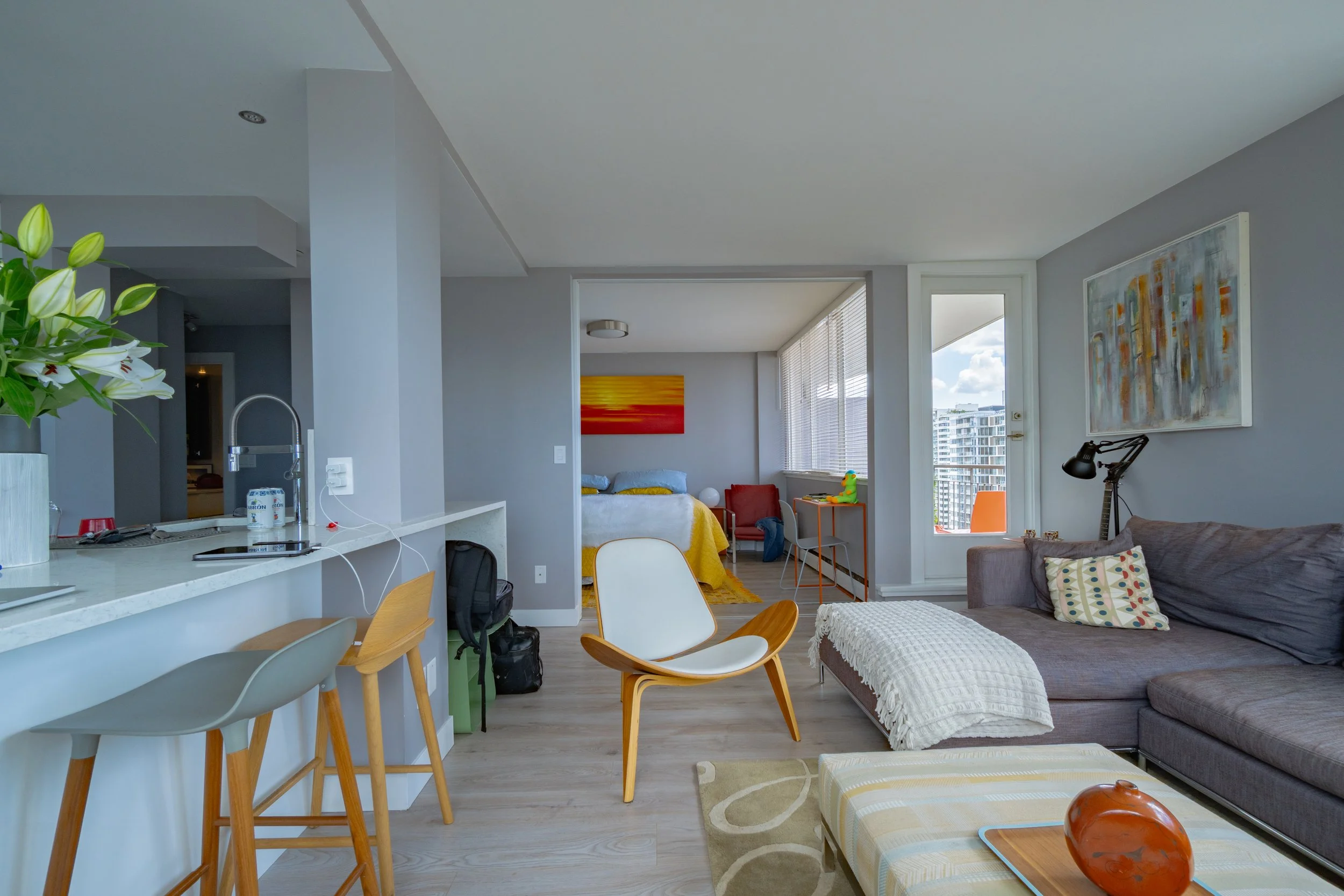 Open-concept living room and kitchen area in a modern apartment, with a gray sofa, colorful artwork, a white and yellow ottoman, and large windows with city view. - Vancouver BC, 1903 Comox St.