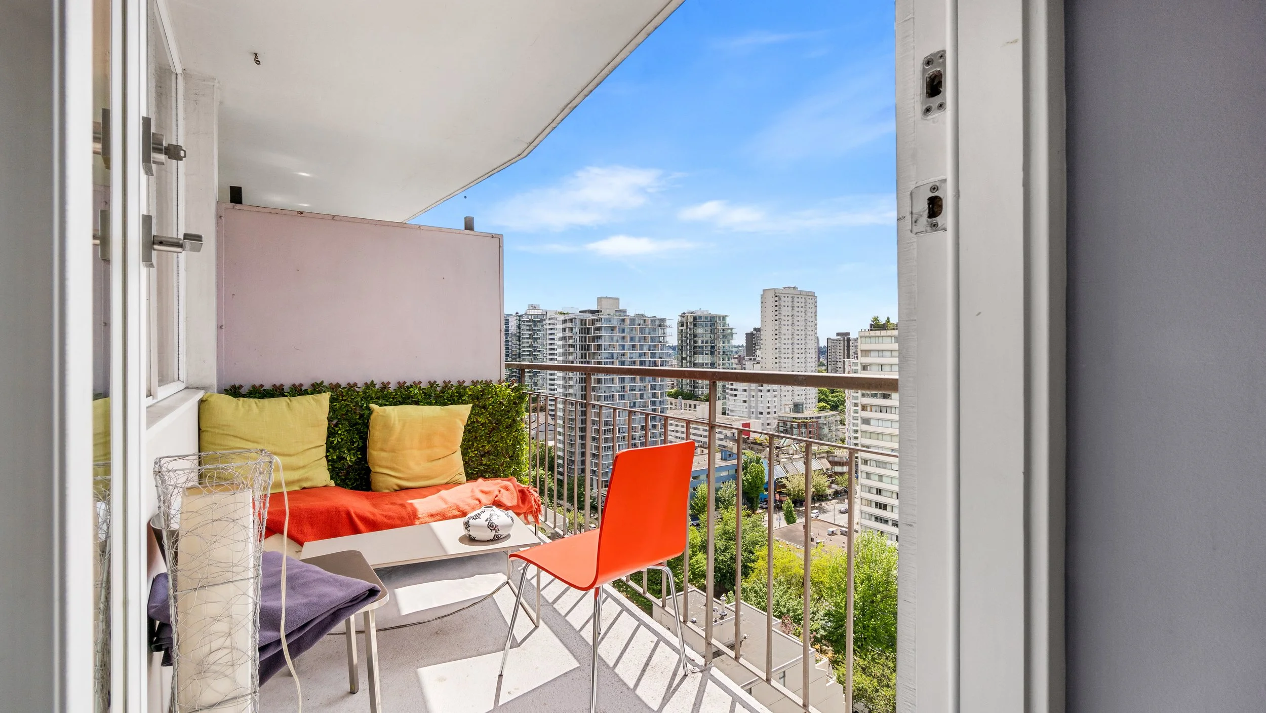 Balcony with colorful cushions, orange and purple chairs, a small table with a decorative object, overlooking a cityscape with high-rise buildings under a blue sky.