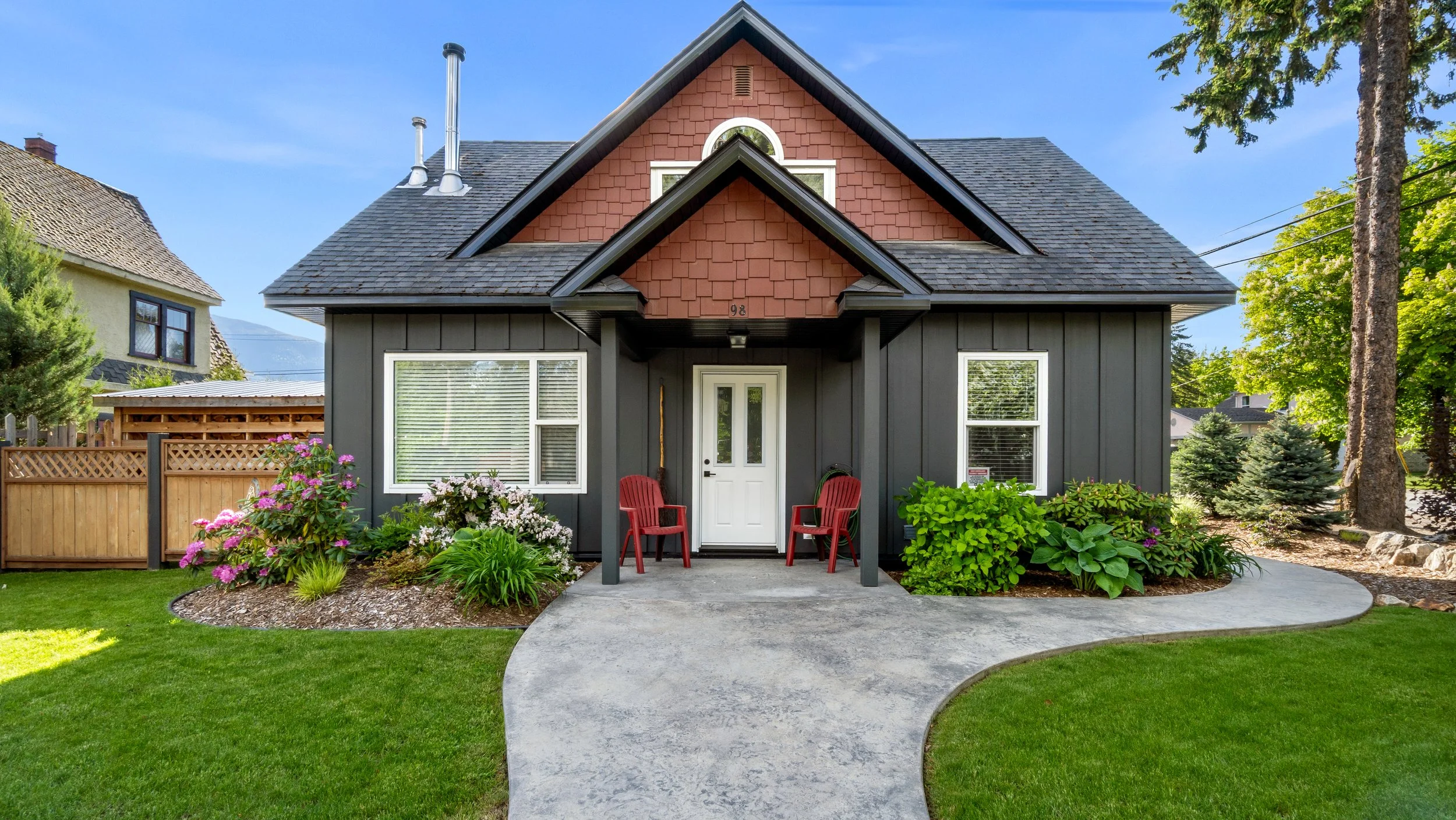 Front view of a dark gray house with red shingles on the gable, white window frames, and a white door. Two red chairs sit on the porch, surrounded by lush green bushes and colorful flowers. A curved concrete pathway leads to the entrance, with a lawn and tall trees in the background.