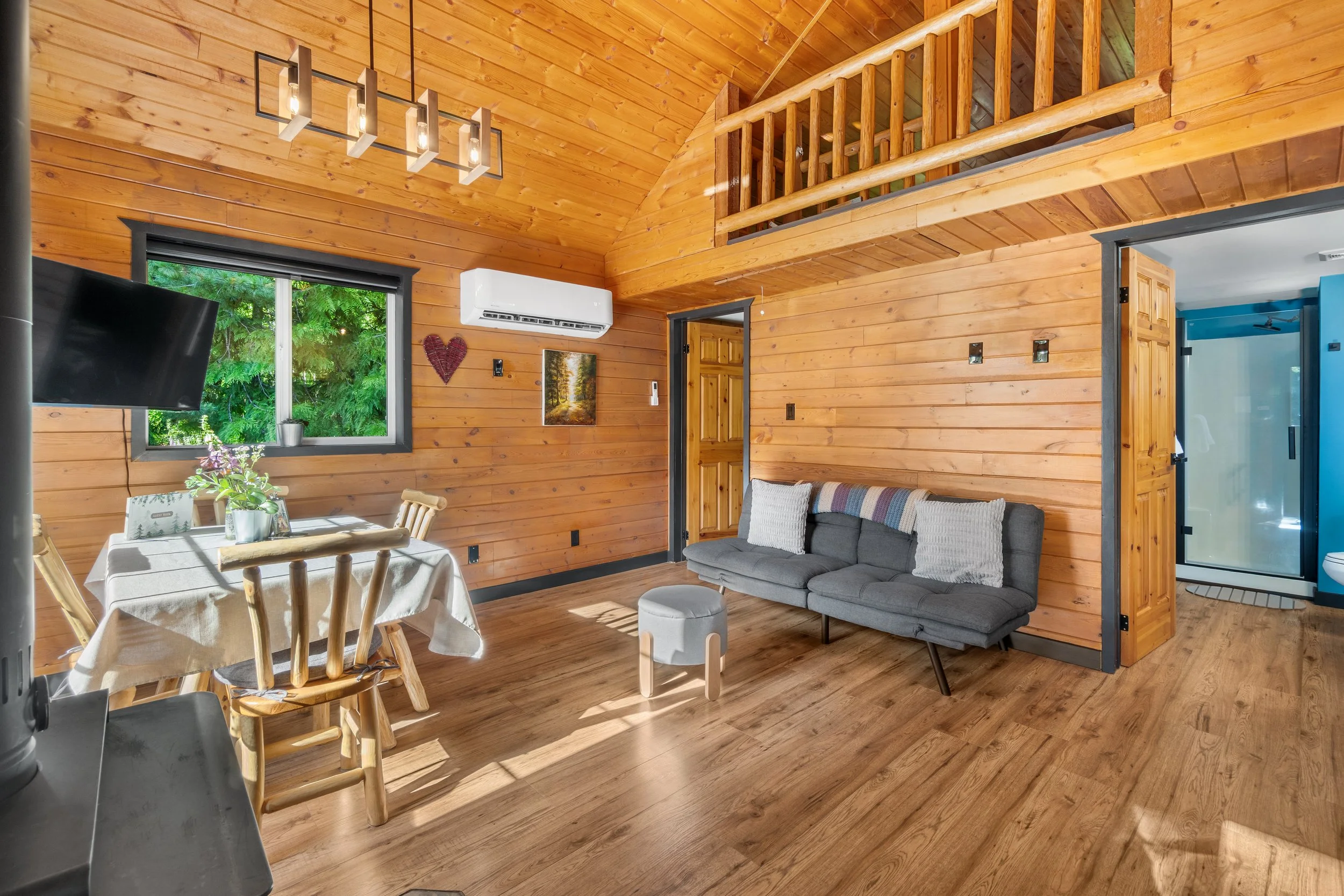 Interior view of a cozy log cabin living room with wooden walls and ceiling, a gray sofa with pillows, a wooden dining table with chairs, a mounted flat-screen TV, a window showing greenery outside, and a loft area with wooden railing.