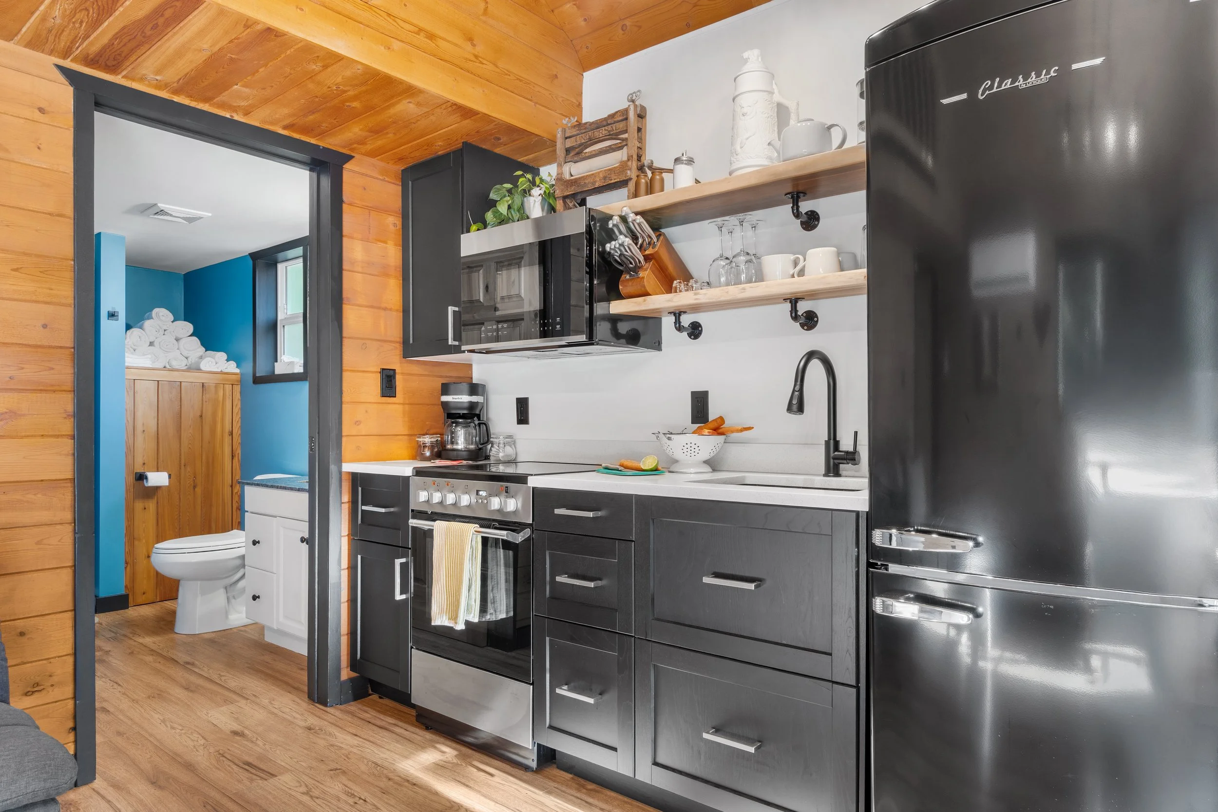 Interior of a rustic kitchen with black cabinets, open wooden shelves, stainless steel appliances, and a wood-paneled ceiling. There are various kitchen items like glasses, cups, teapot, and a coffee maker.