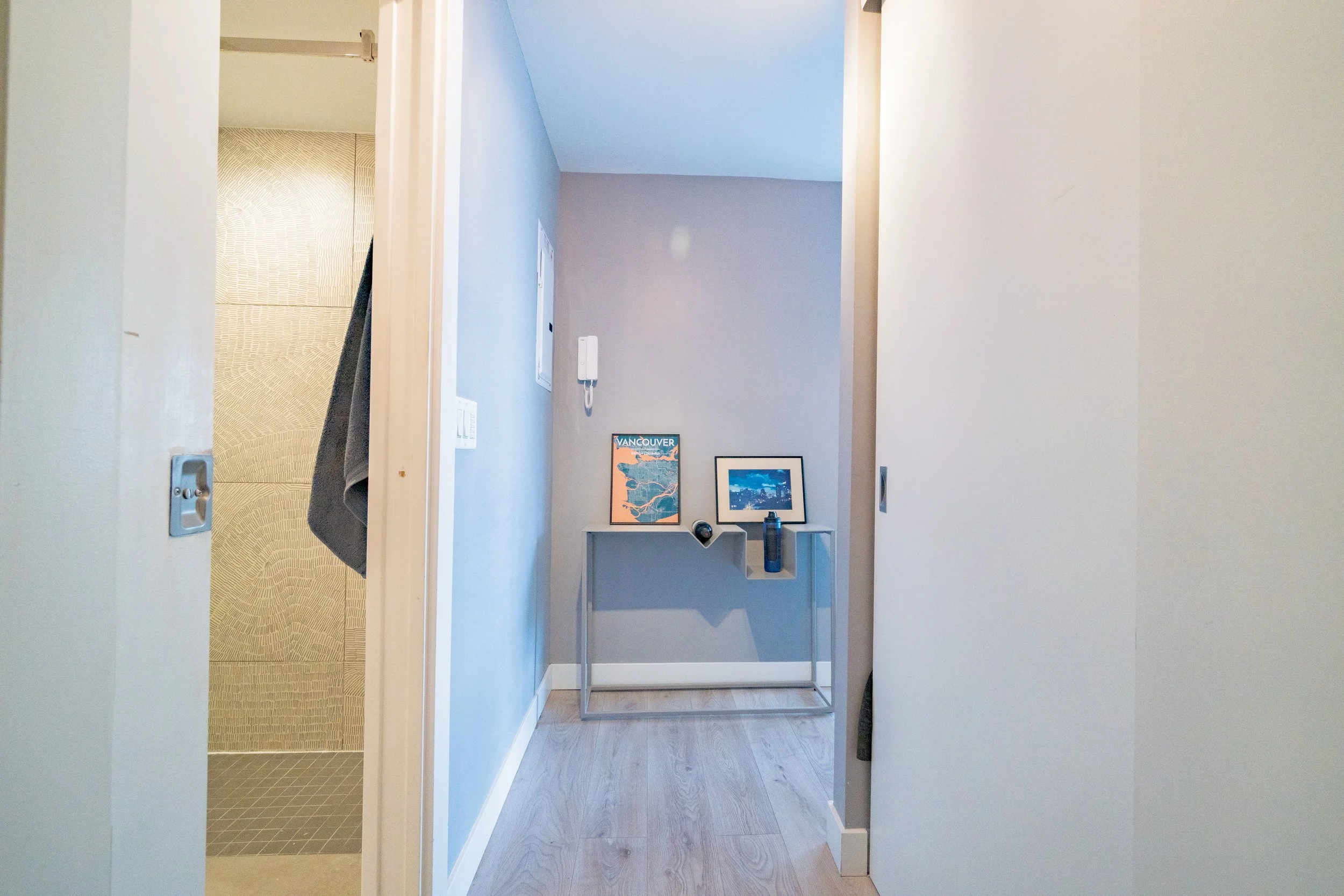 Interior view of a hallway with a small table holding framed pictures, a speaker, and a water bottle. Walls are painted blue and gray, with a door open to a room with beige patterned wallpaper. On the wall, there is an intercom system and light switches.