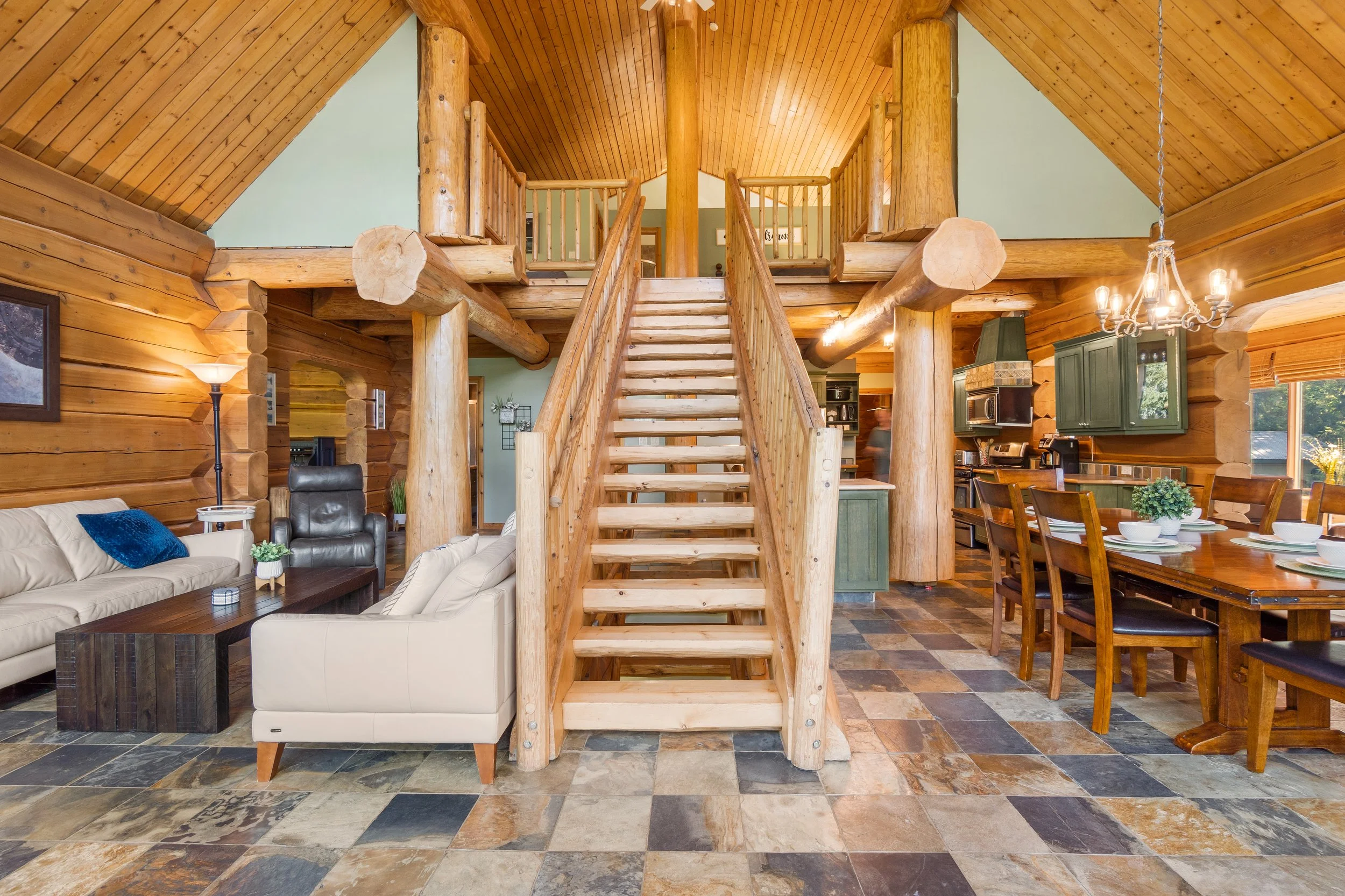 Interior view of a rustic log cabin living and dining area with wood ceiling, staircase, sofa, chairs, and kitchen.