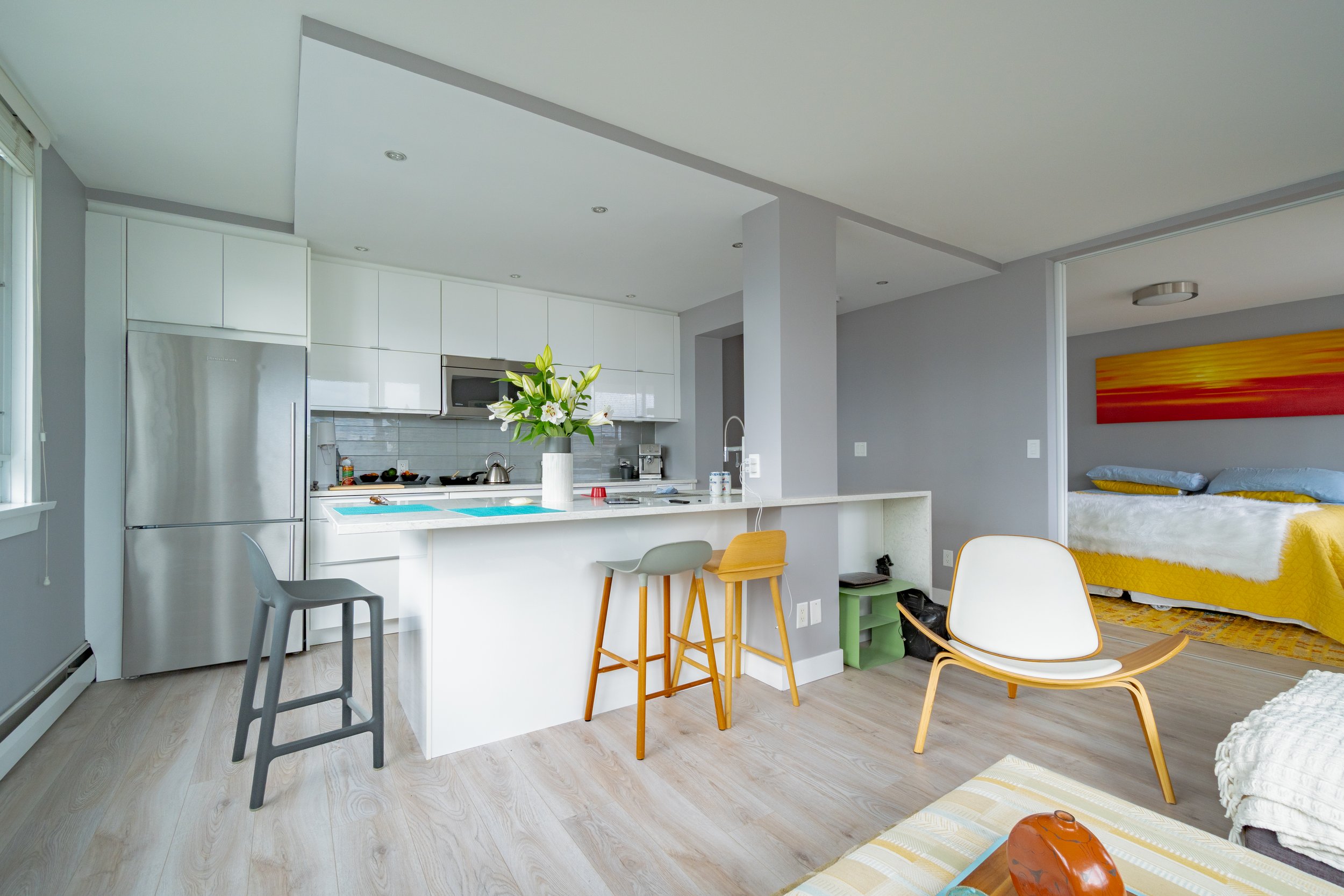 Open-concept living space combining a kitchen and bedroom with modern decor, light wooden flooring, white cabinets, gray walls, and yellow accents. Vancouver BC, 1903 Comox St.