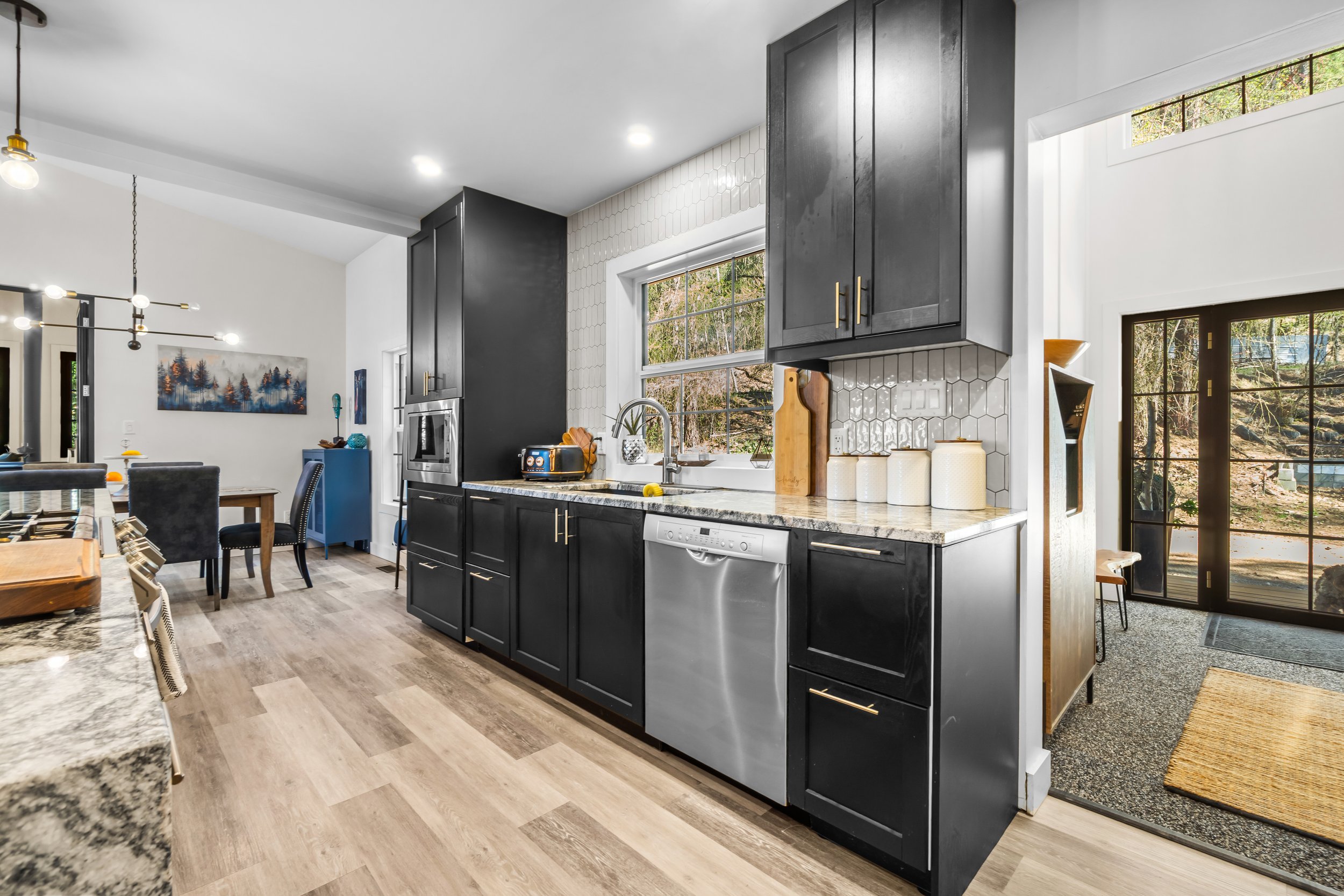 Modern kitchen with black cabinets, white hex tile backsplash, stainless steel dishwasher, and a window overlooking a wooded outdoor area.