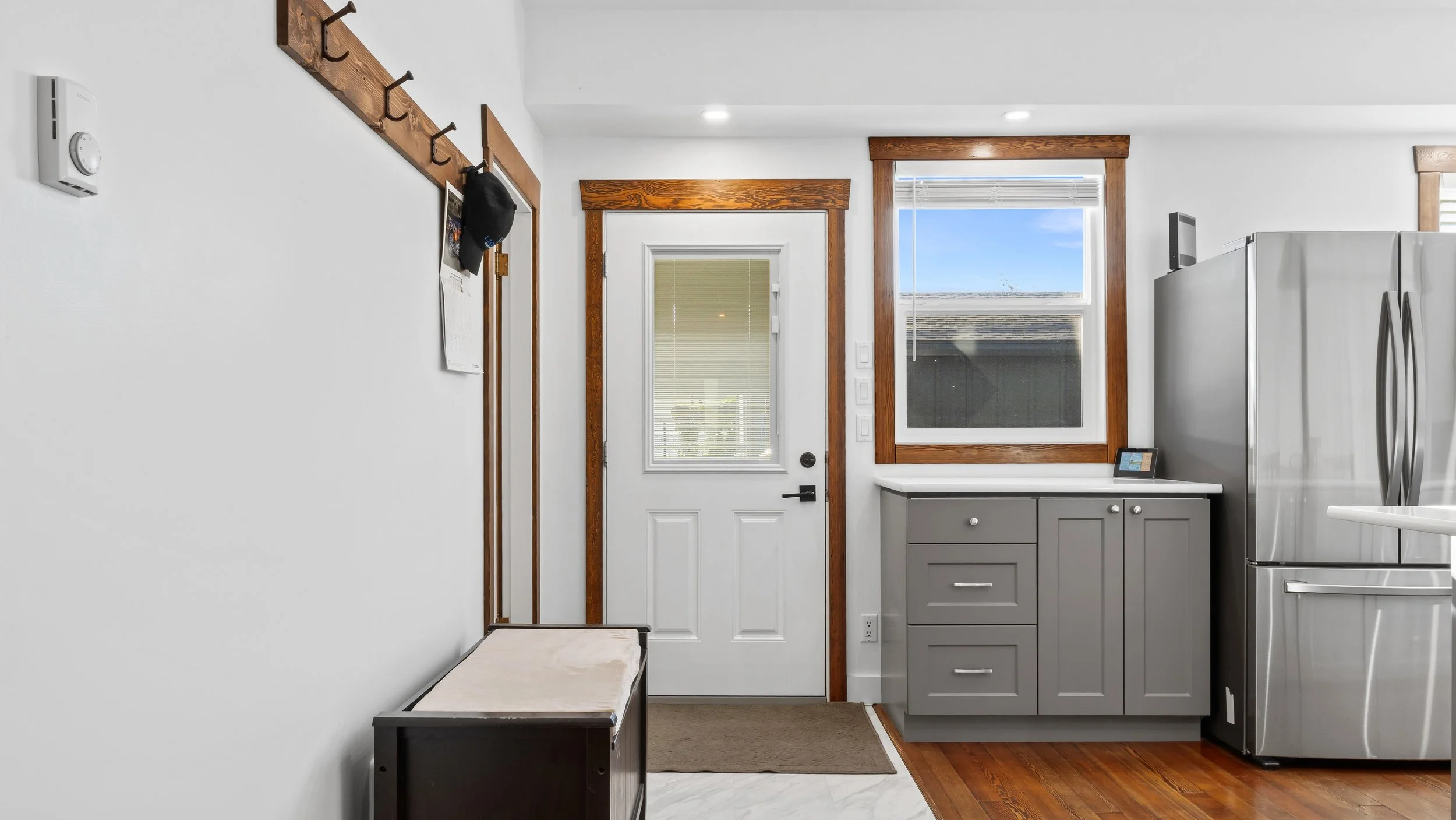 Interior view of kitchen entryway with white walls, wooden trim, a white door with a window and blinds, a gray cabinet, a stainless steel refrigerator, a small tablet on the cabinet, and a bench at the left has a white cushion. Nakusp, 98 2nd Ave