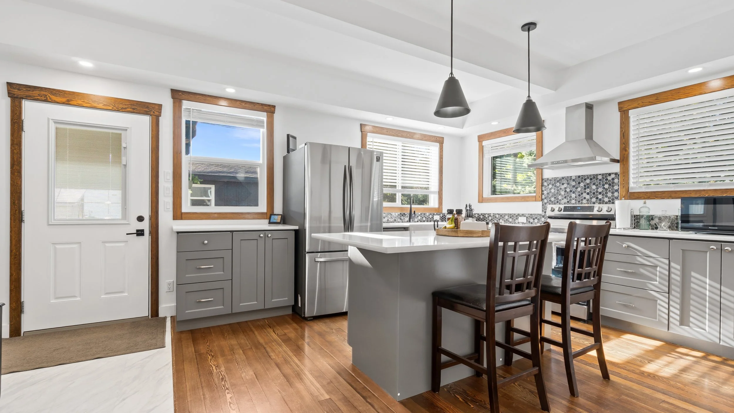 Kitchen with white cabinets, gray drawers, stainless steel refrigerator, white countertop, gray backsplash, window with blinds, wooden trim, black pendant lights, stove, microwave, and dining chairs.