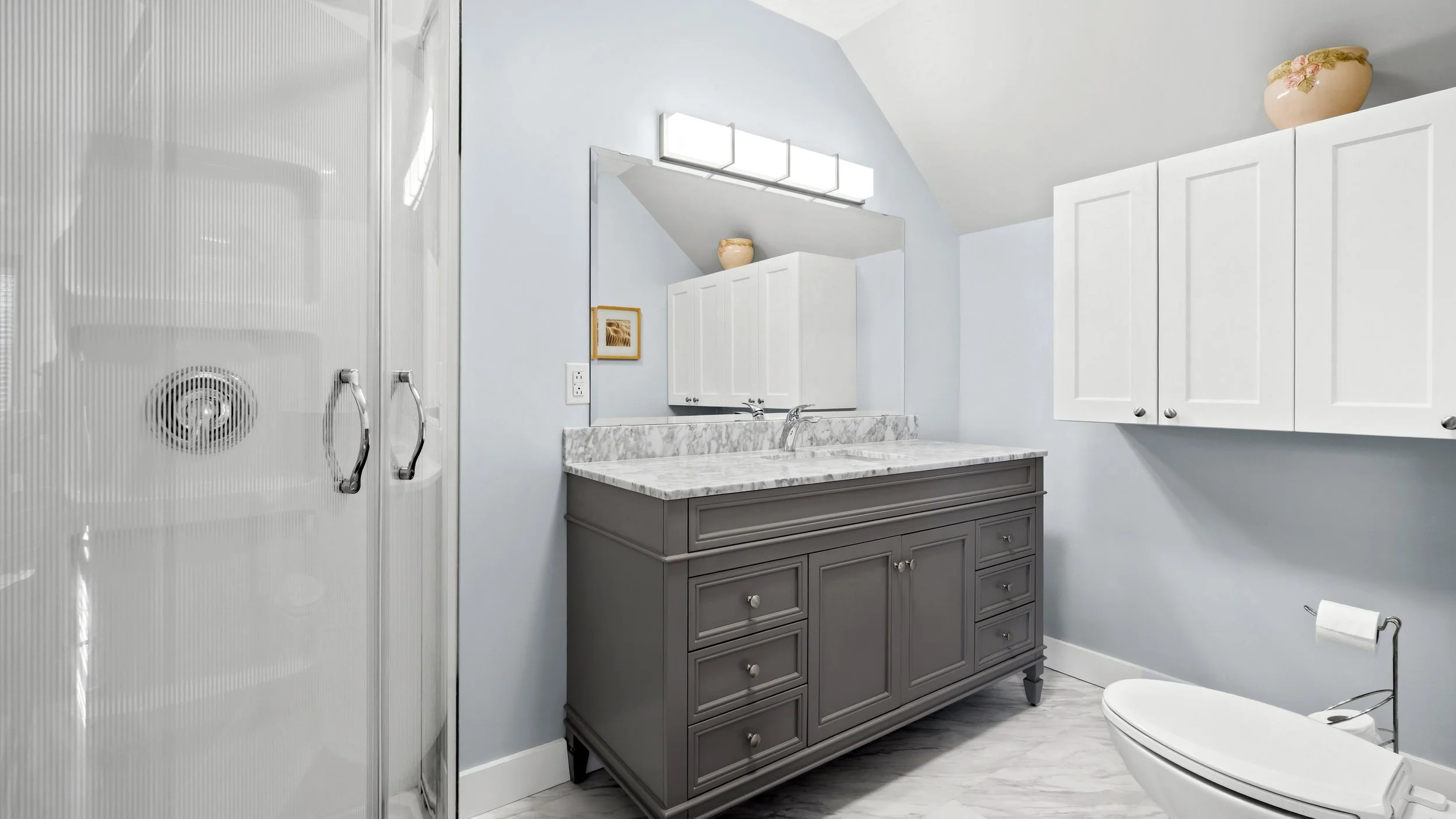 A modern bathroom with a gray vanity featuring a marble countertop, white cabinets above, mirror, and a glass shower door on the left. The bathroom has light blue walls, a framed art, and decorative vases on top of the cabinets. - Nakusp, 98 2nd Ave