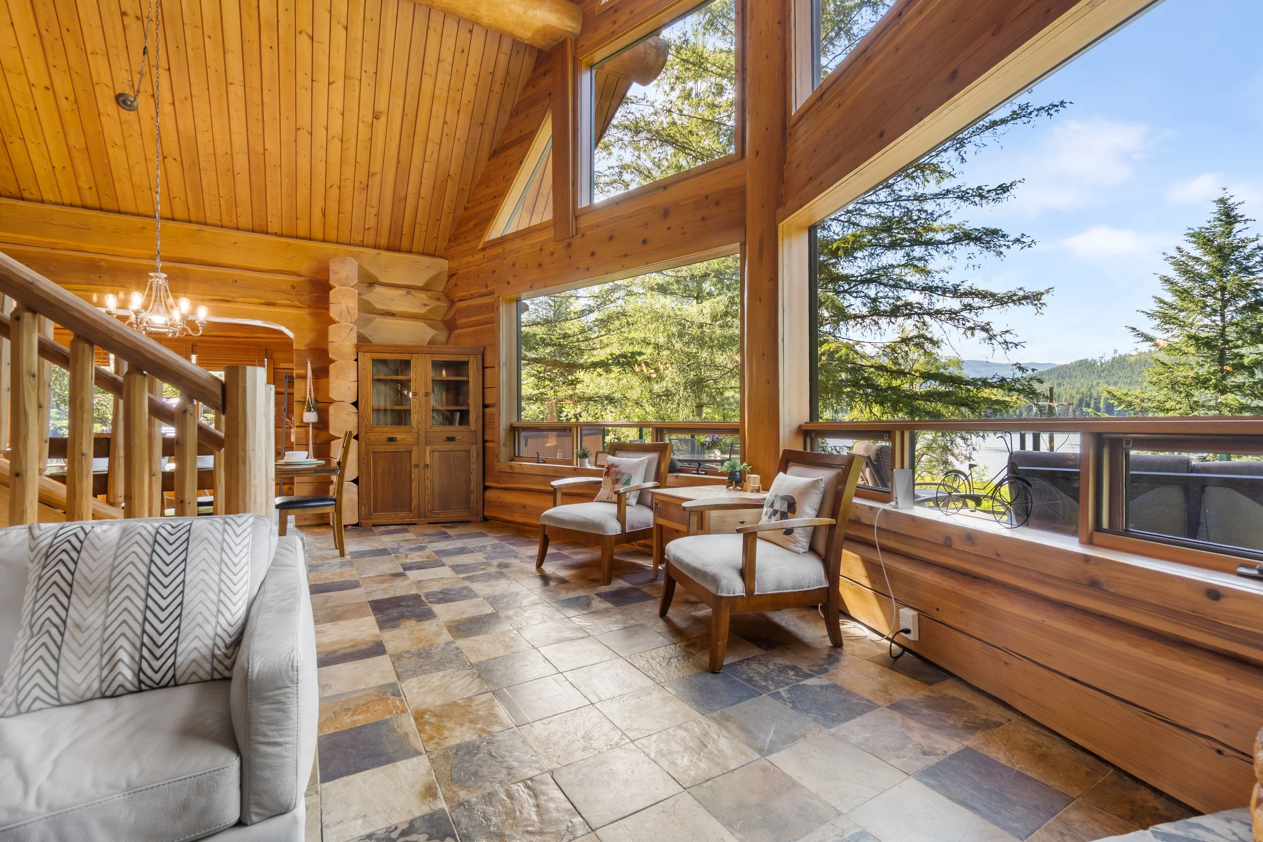 Interior view of a wooden cabin living room with large windows overlooking trees and mountains, furnished with a white sofa, wooden chairs with cushions, a wooden cabinet, and a chandelier.