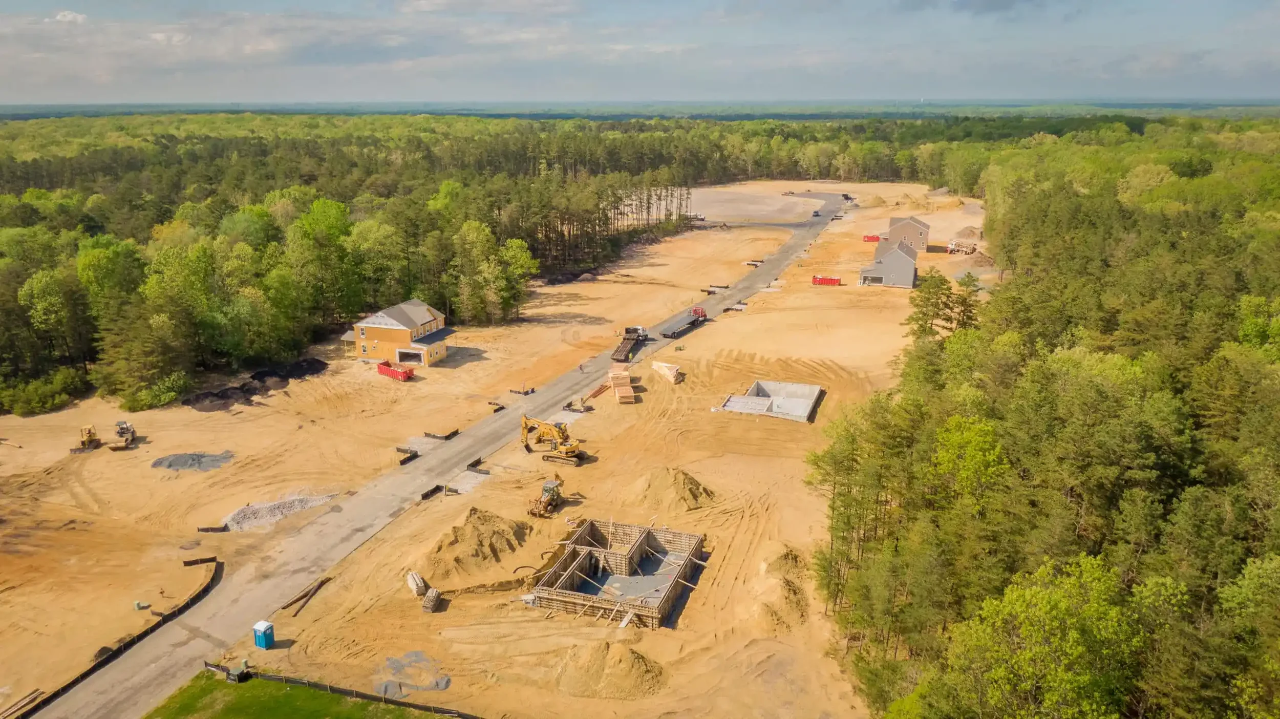 Aerial view of a construction site in a wooded area with houses under construction, trucks, and heavy machinery on dirt and gravel road, surrounded by green trees.