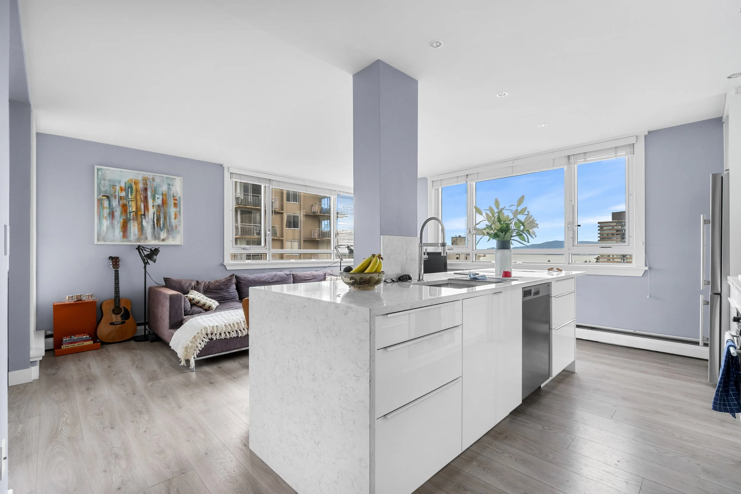 A modern open-concept living room and kitchen with large windows showing a city view. The room features a purple sofa, a guitar, a bookend, a lamp, a painting, and a banana bowl on the kitchen island. The kitchen has white cabinets and a marble countertop.